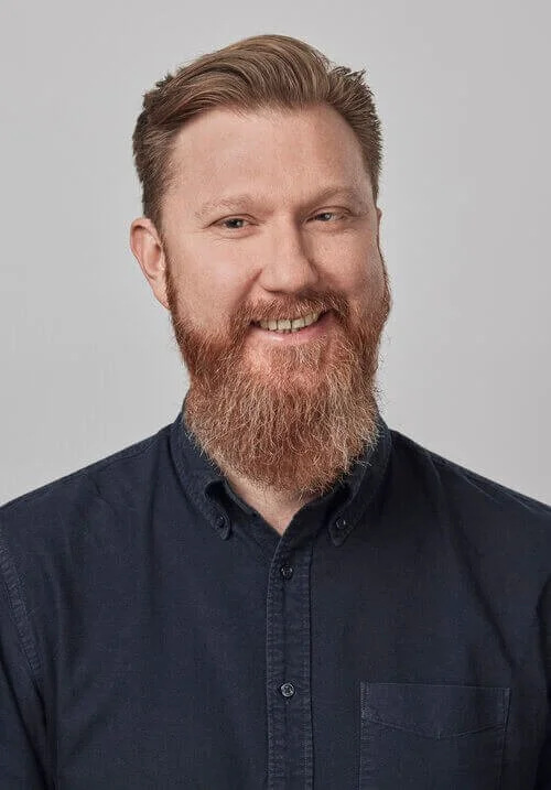 Smiling man with beard and short hair in dark shirt against light gray background.