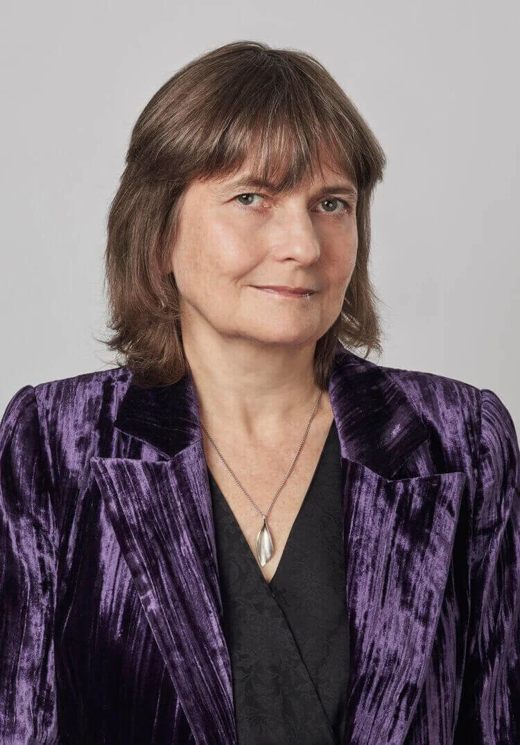 Woman in purple velvet blazer and black top with silver pendant, on light gray background.