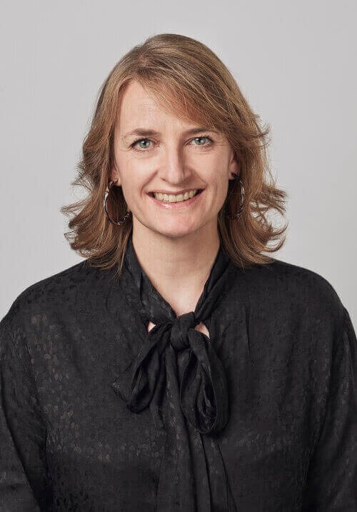 Woman with light brown hair in black blouse with bow, smiling against light gray background.