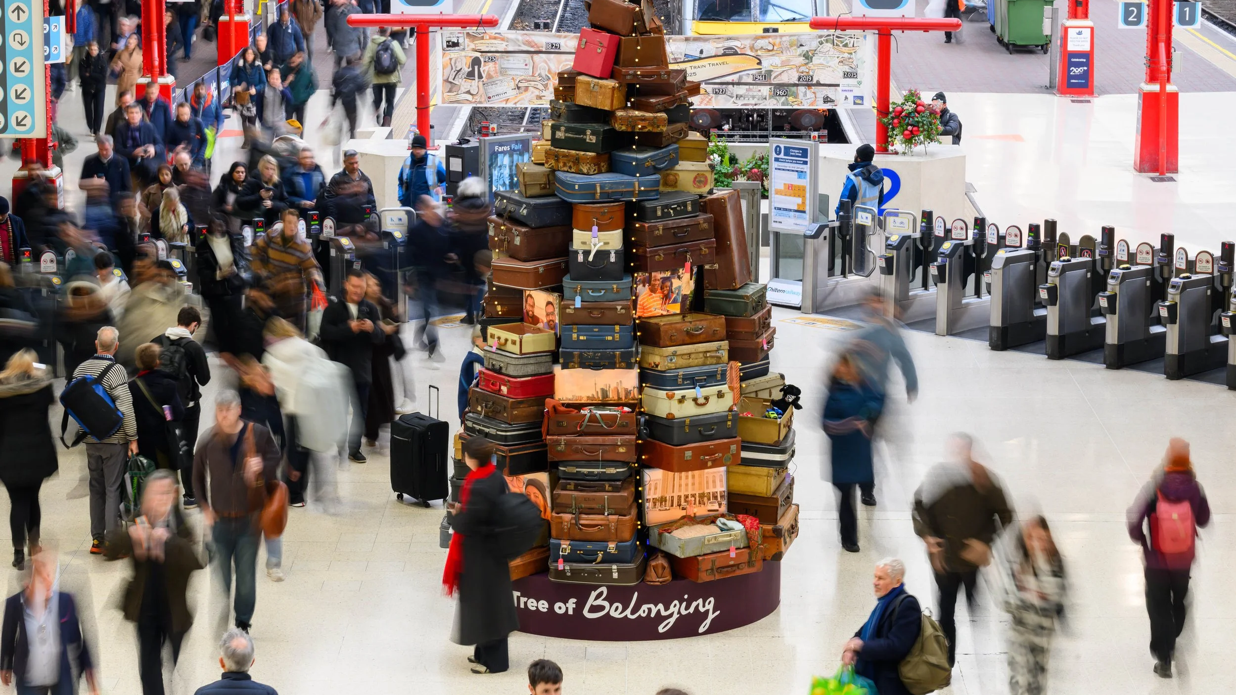 A tall Christmas tree made from stacked vintage suitcases stands in a busy train station, with blurred commuters moving around it.