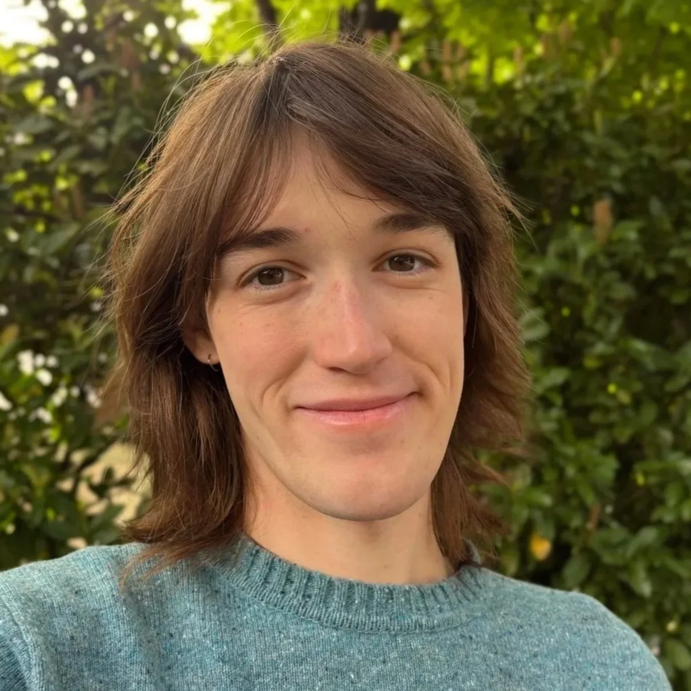 A young adult with medium length brown hair smiles softly into the camera, greenery in the background.