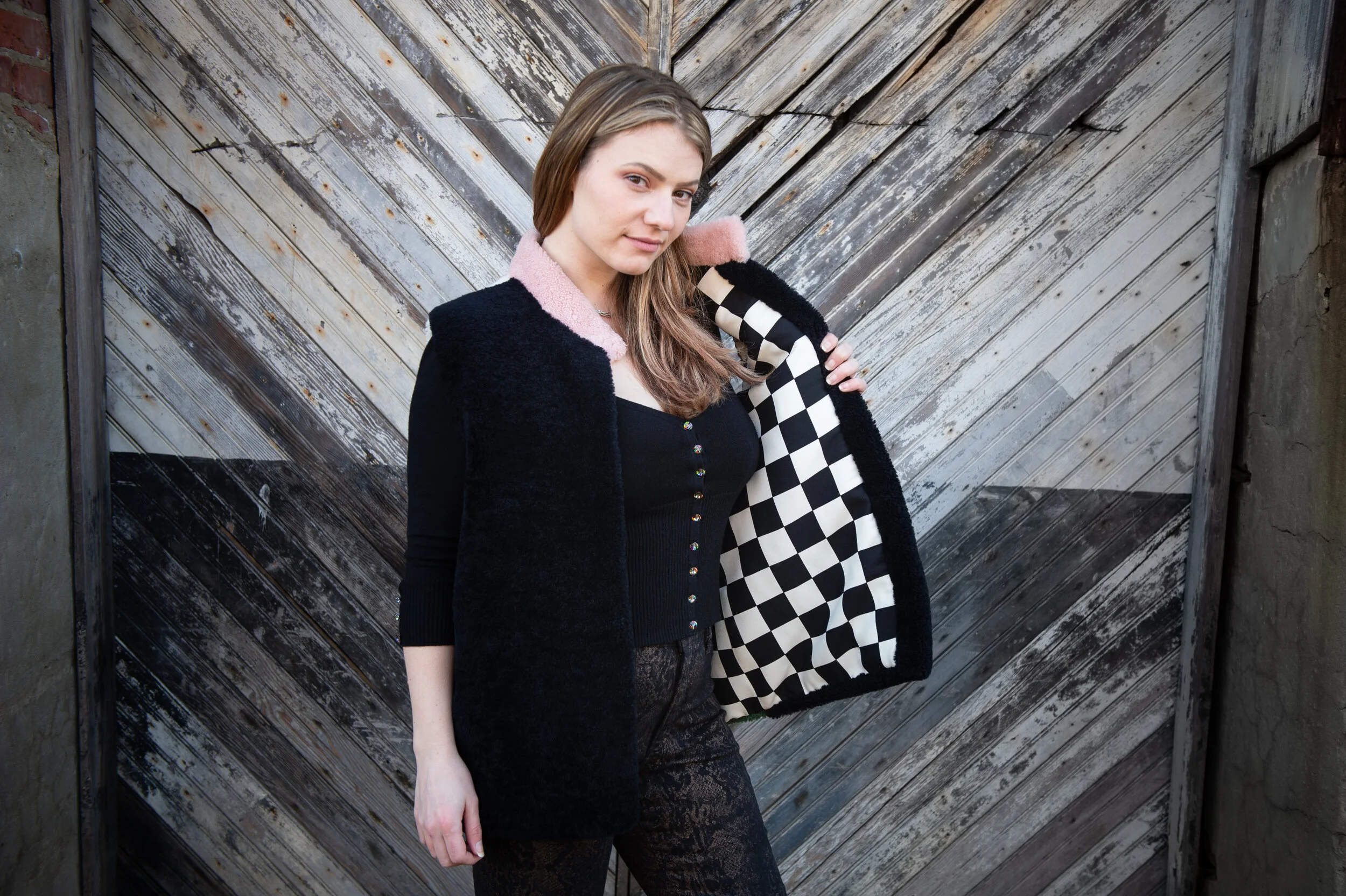 A young woman with light skin and long light brown hair standing in front of a weathered wooden barn door with diagonal planks. She is wearing a black long-sleeve top, a navy blue cardigan with multicolored buttons, and a black fuzzy jacket with pink