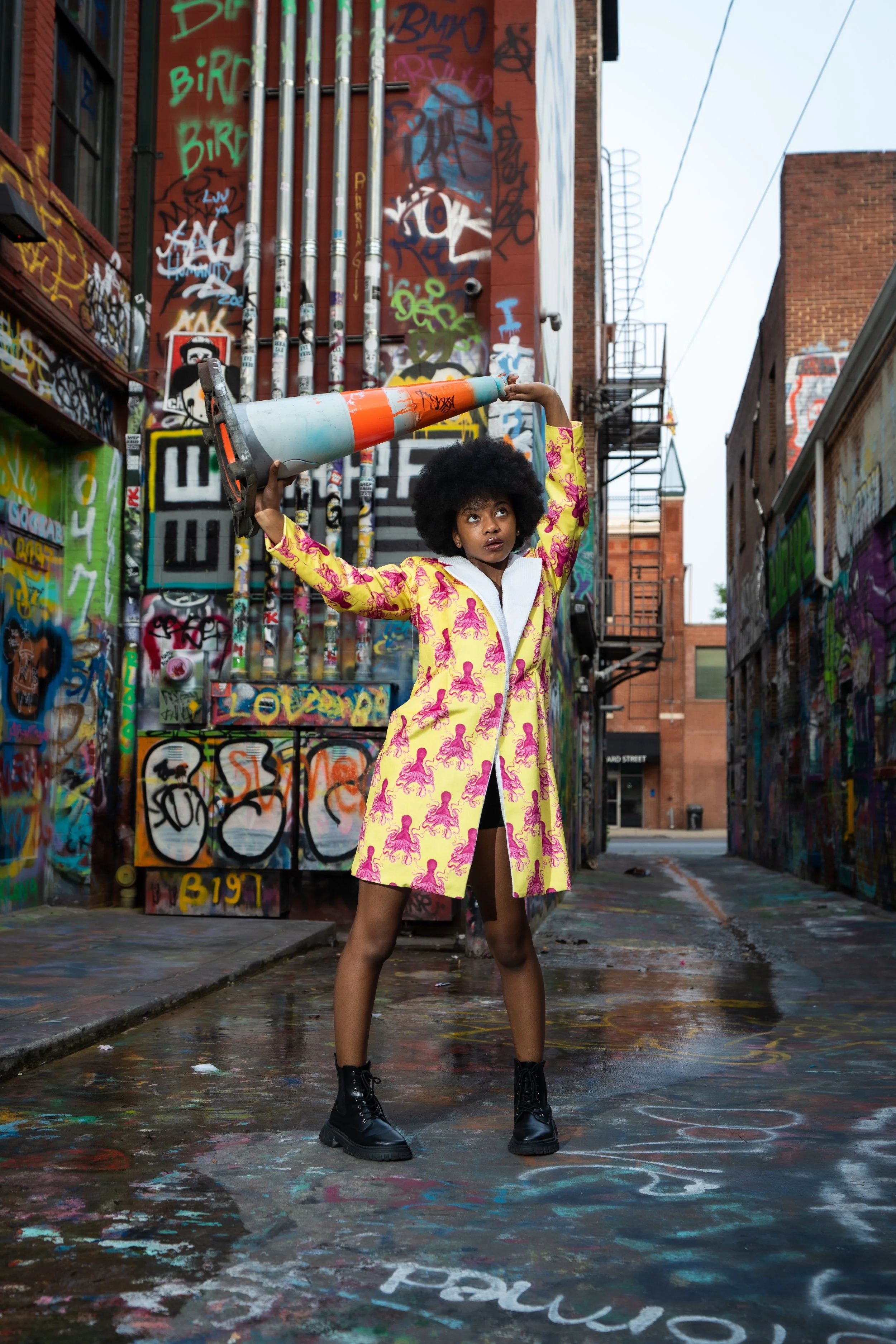 A young woman with curly hair wearing a colorful yellow coat with pink patterns and black combat boots stands in an alley covered in graffiti. She is holding a large traffic cone above her head and looking upward.
