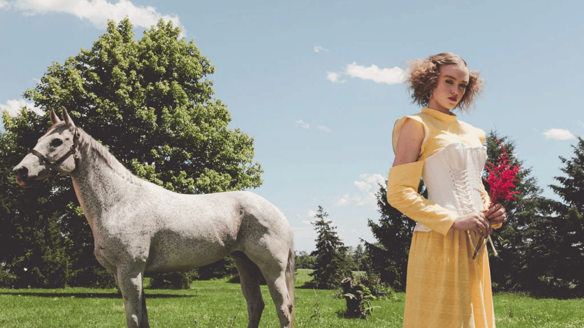A woman with curly hair in a yellow dress holding a bunch of pink flowers standing beside a white horse in a park with green trees and a blue sky.