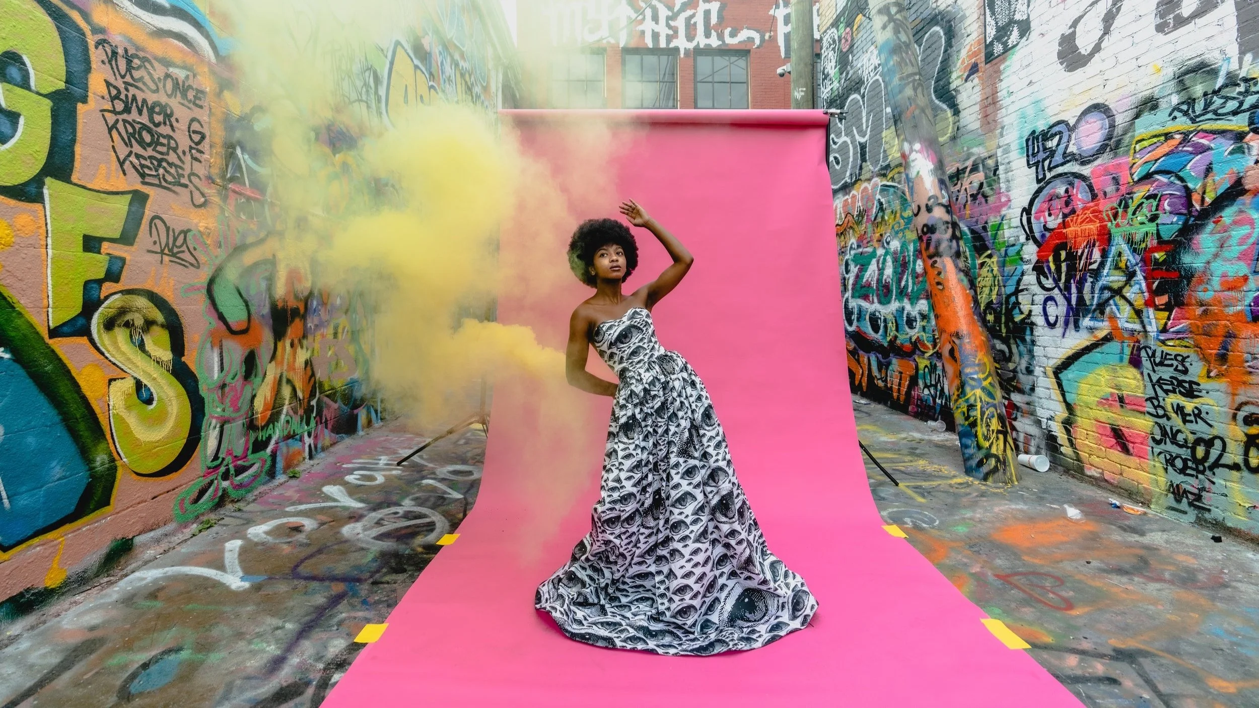 A woman in a black and white patterned dress poses on a pink backdrop in a graffiti-covered alley, with yellow smoke in the air.