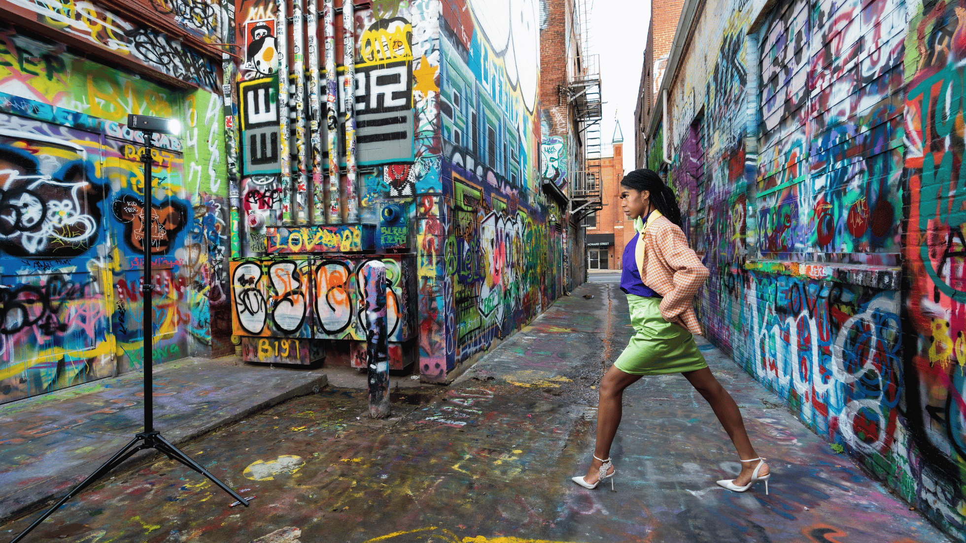 A woman in a colorful outfit, with a violet top and a bright green skirt, posing with her hands in her pockets in a narrow alleyway covered in vibrant graffiti art on the walls and ground. There is a fire escape on one side and a street sign reading "Howard Street" in the background.