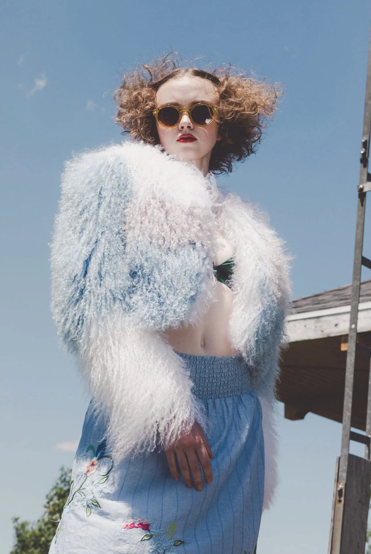 A woman with curly hair wearing sunglasses, a white fuzzy jacket, and floral shorts stands outdoors against a clear blue sky.