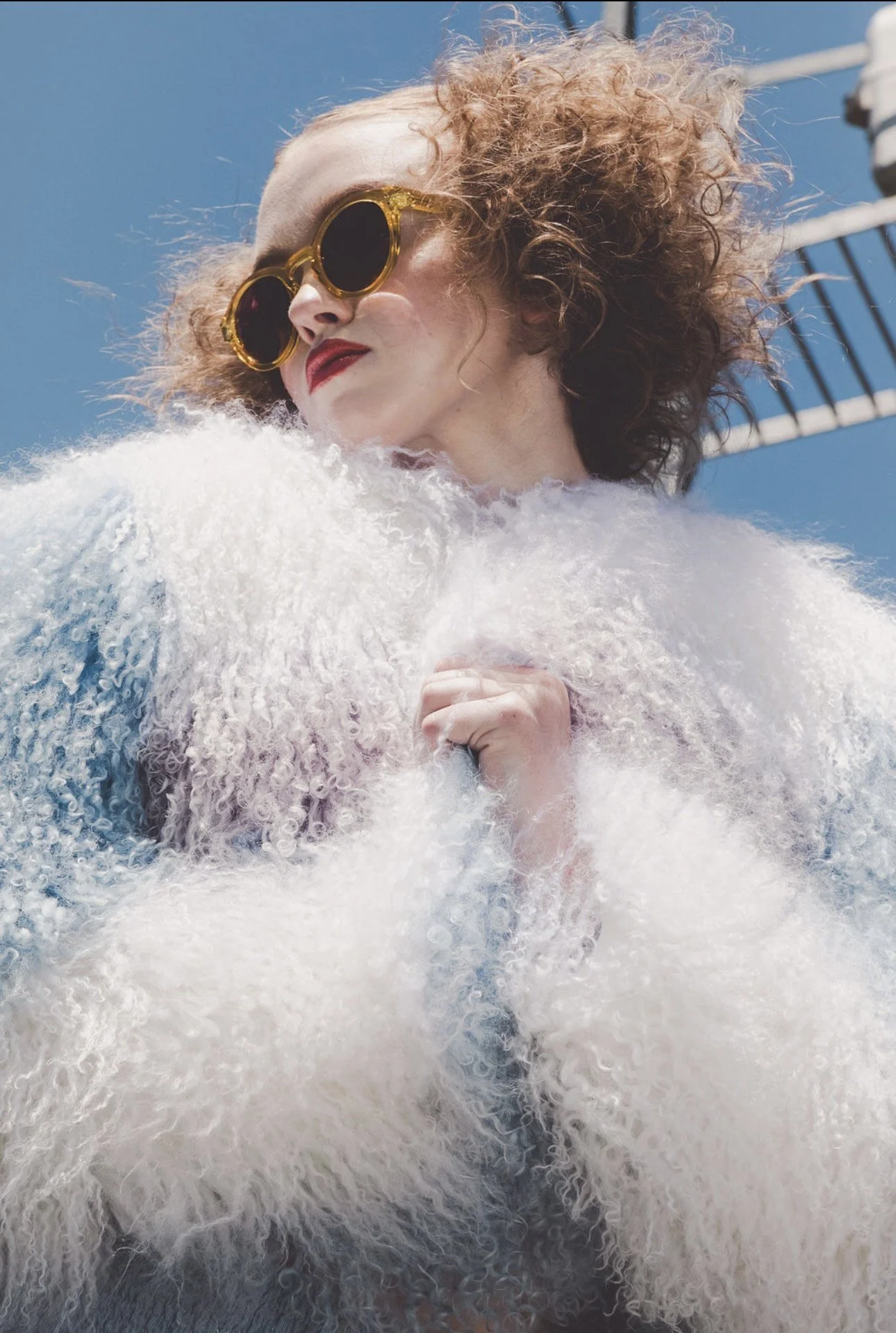 A woman with curly red hair and red lipstick wearing sunglasses and a white fluffy coat, standing outdoors under a clear blue sky.