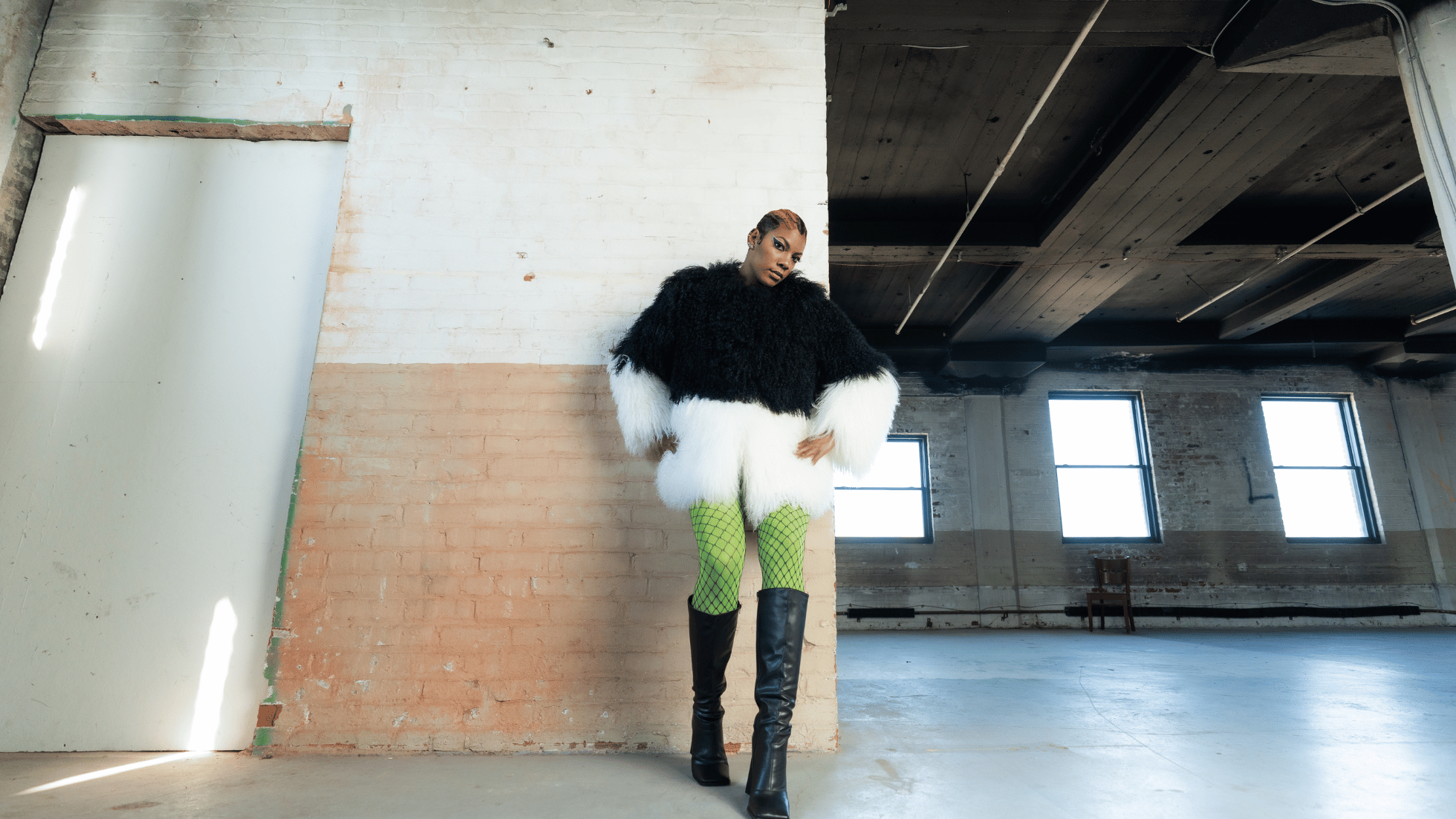 A woman standing in an industrial-style room with exposed brick walls and large windows, wearing a black and white fur coat, neon green fishnet stockings, and tall black boots.