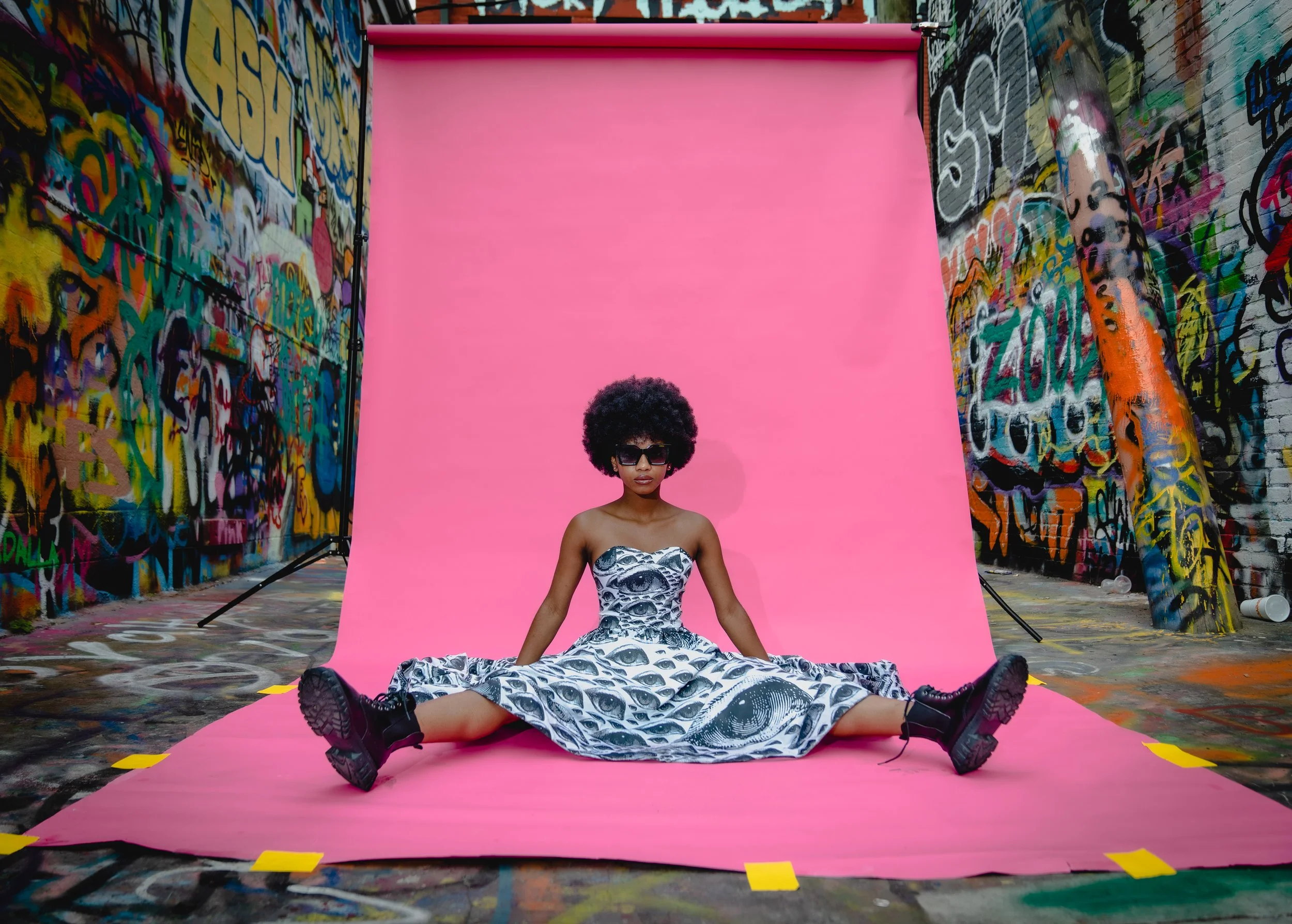 A woman with a large afro hairstyle and sunglasses sits on a pink backdrop with her legs spread apart, in front of colorful graffiti-covered walls.