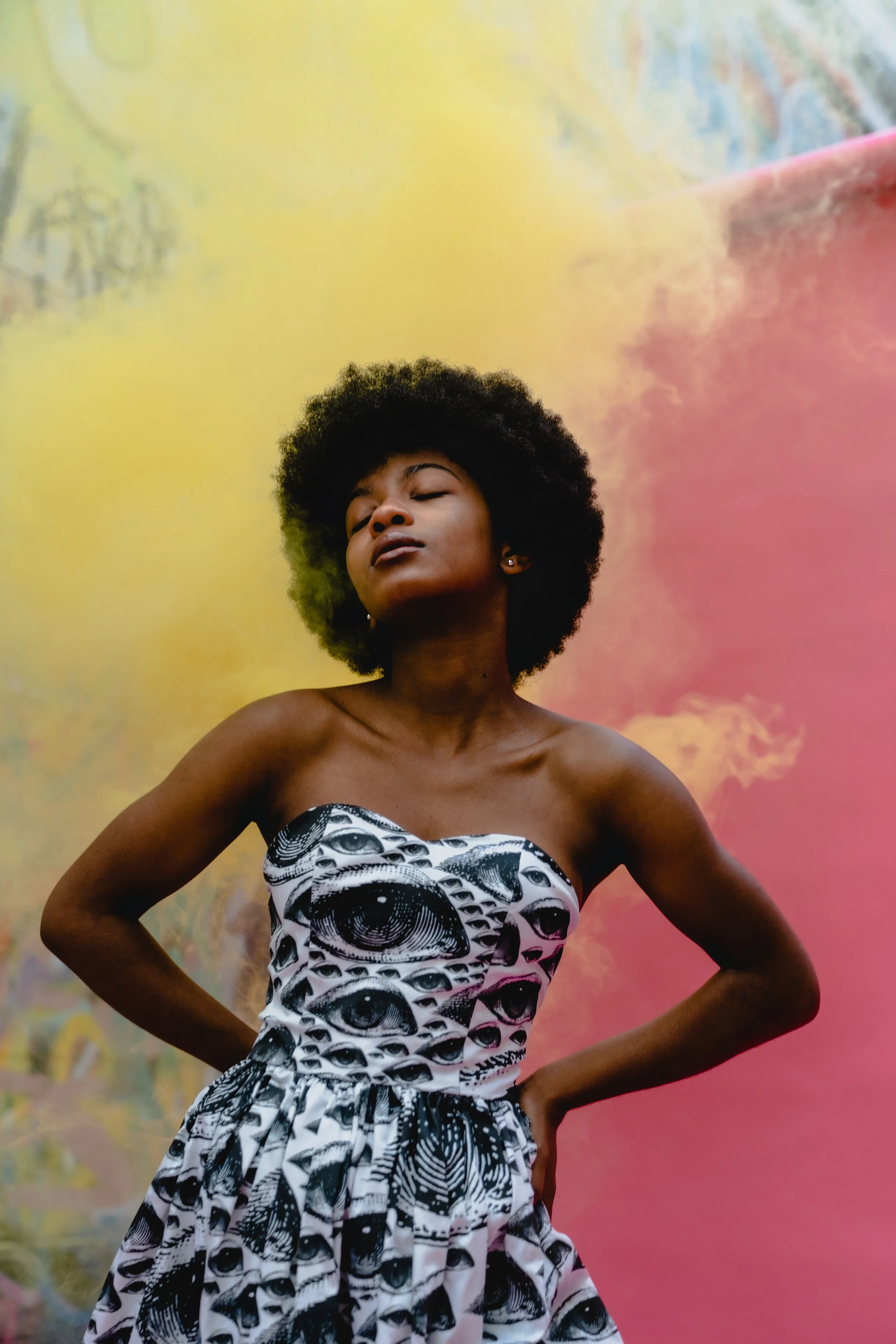 A woman with dark skin and an afro hairstyle posing with her hands on her hips, wearing a strapless dress with an eye pattern, standing against a colorful, abstract background.
