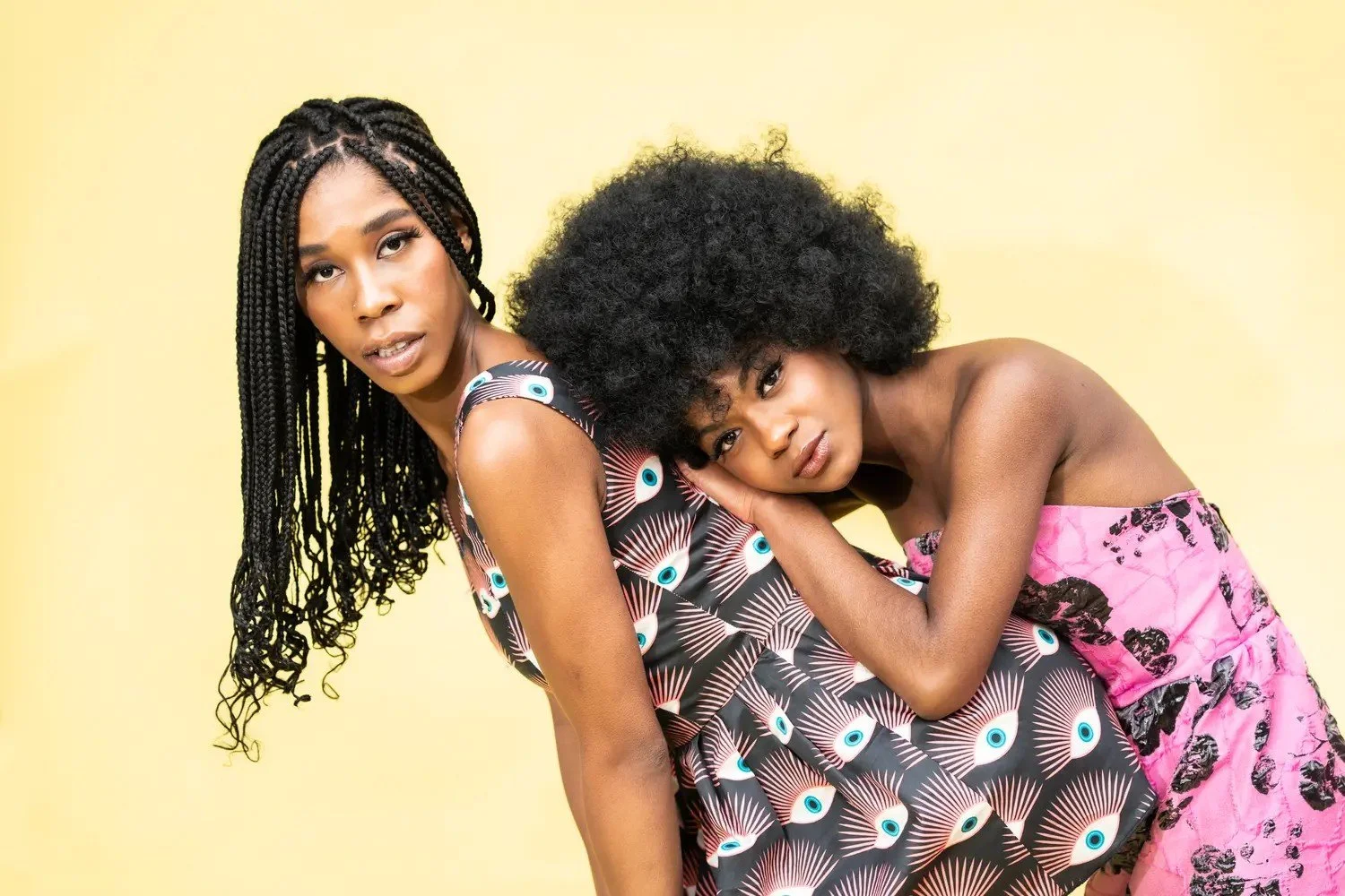 Two women with dark curly hair against a yellow background, one with long braided hair and the other with a voluminous afro, pose closely with a look of concern or contemplation.