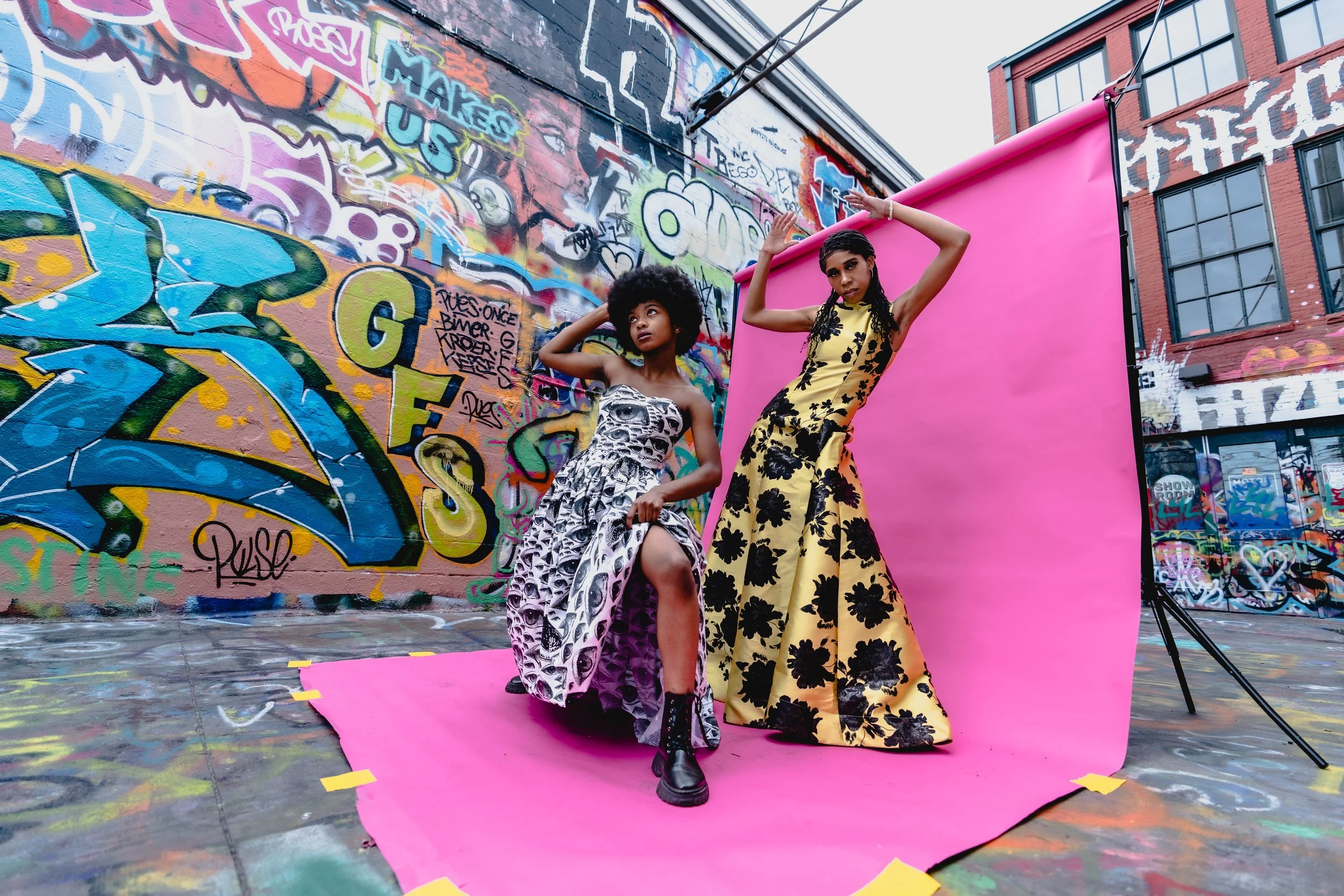 Two women posing on a pink photography backdrop against a graffiti-covered wall in an outdoor urban alley.