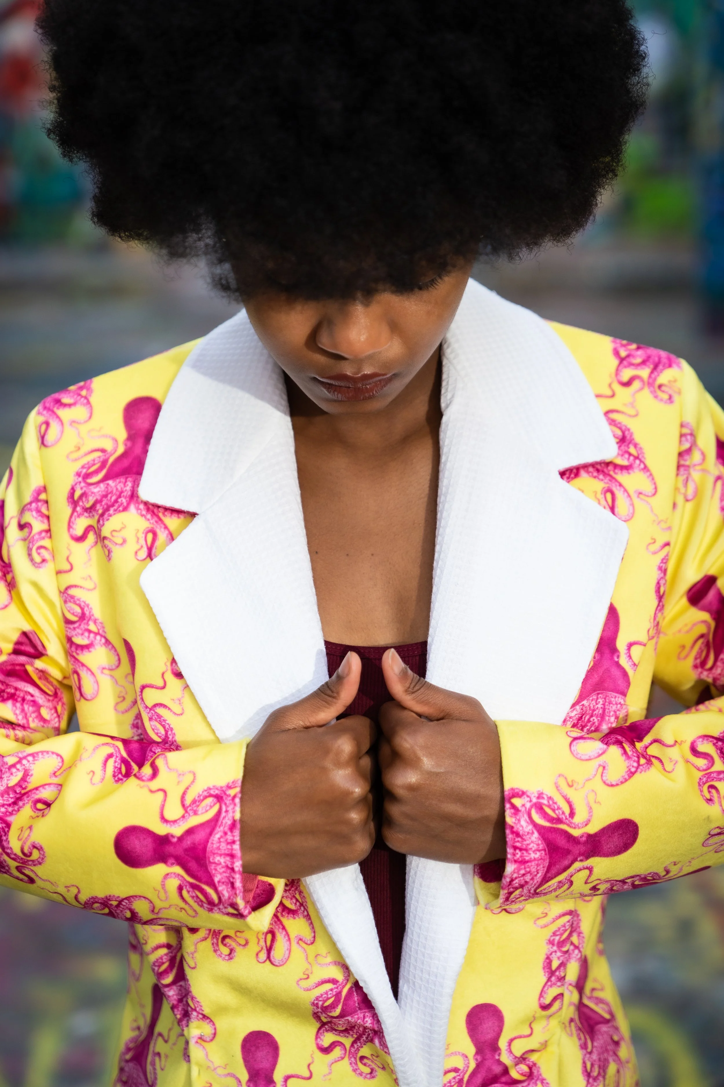 A woman with an afro hairstyle, wearing a yellow jacket with pink tentacle patterns, a white collared shirt, and a dark top underneath. She is looking down with her hands clasped together in front of her, in a contemplative pose.