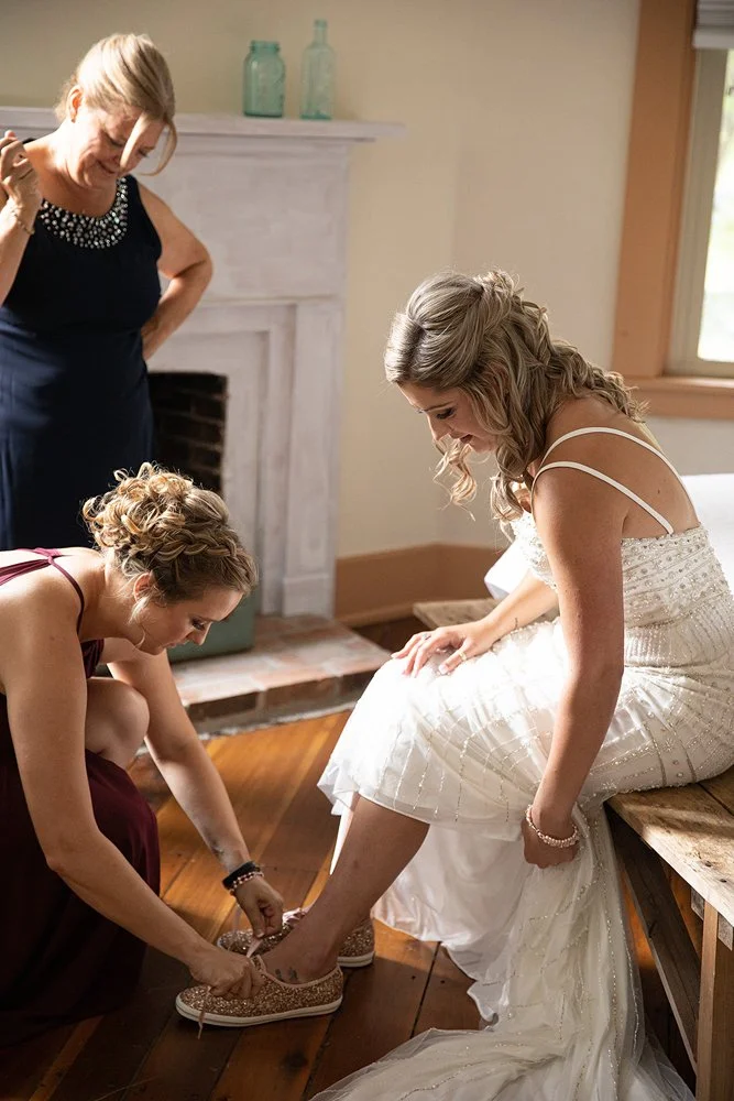 Bride sitting on a bench with a long white wedding dress, as a woman in a purple dress helps her put on sparkly sneakers, while another woman in a dark dress observes in a room with hardwood floors.