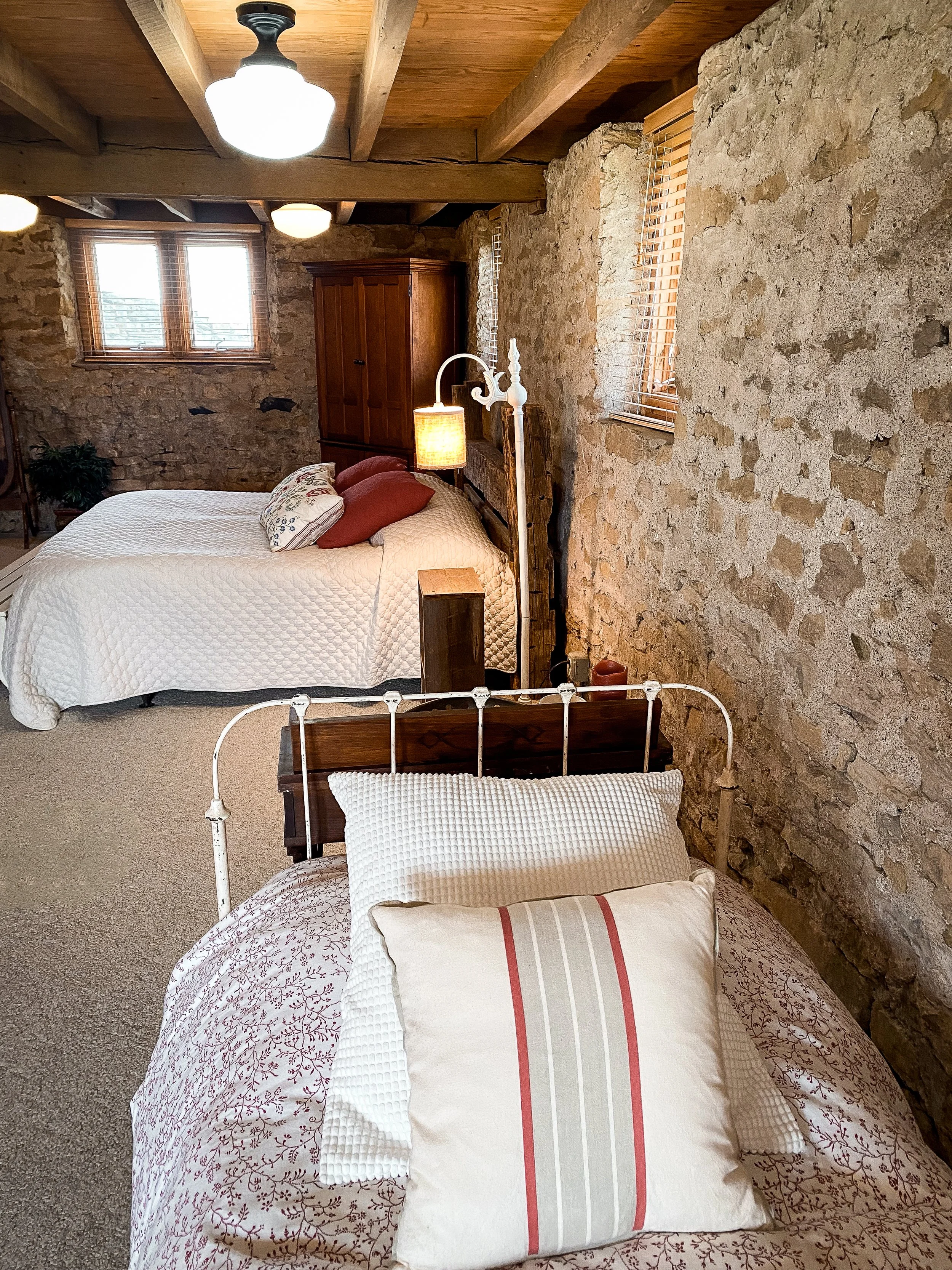Rustic bedroom with stone walls, two beds with white bedding, wooden ceiling with exposed beams, carpeted floor, and warm lighting. Wedding Retreat. Barn Wedding Venue. Yellow Springs, Ohio