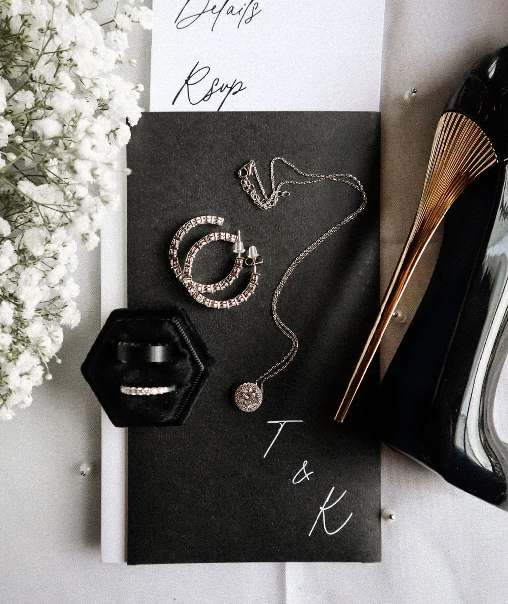 Black high heel shoe, diamond jewelry including a necklace and hoop earrings on a black envelope, tiny white flowers, and an invitation partially showing words 'Details' and 'RSVP'."