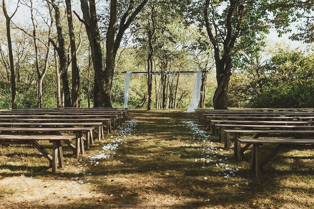 Outdoor wedding venue with wooden benches, an aisle of flower petals, and a rustic wooden arch draped with fabric, surrounded by trees. The Ohio Barn. Springfield, Ohio. Affordable Wedding venue. Columbus, Ohio