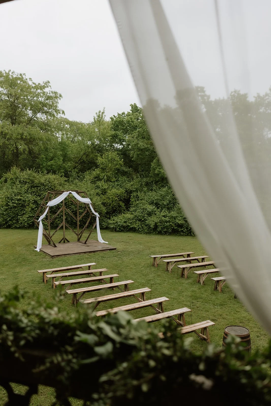 Outdoor wedding venue with wooden benches, a barrel with white flowers, and a geometric arch draped with fabric. Wedding Venue. Ohio Barn Venue. Fairborn, Ohio
