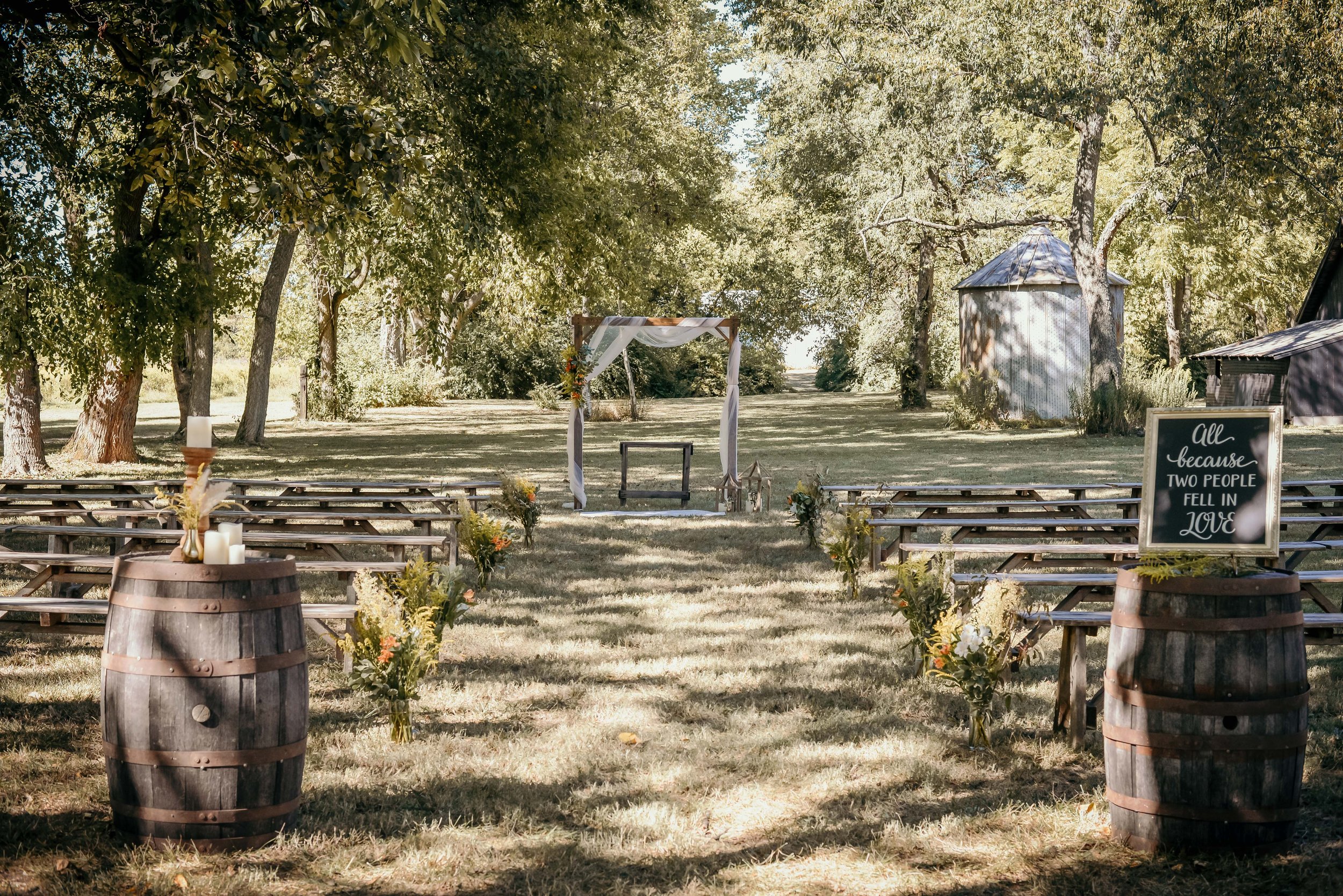 Rustic outdoor wedding ceremony setup with wooden benches arranged in rows facing a decorated archway. The scene includes two barrel tables with candles and a sign reading 'All because two people fell in love.' Surrounded by tall trees and greenery.
