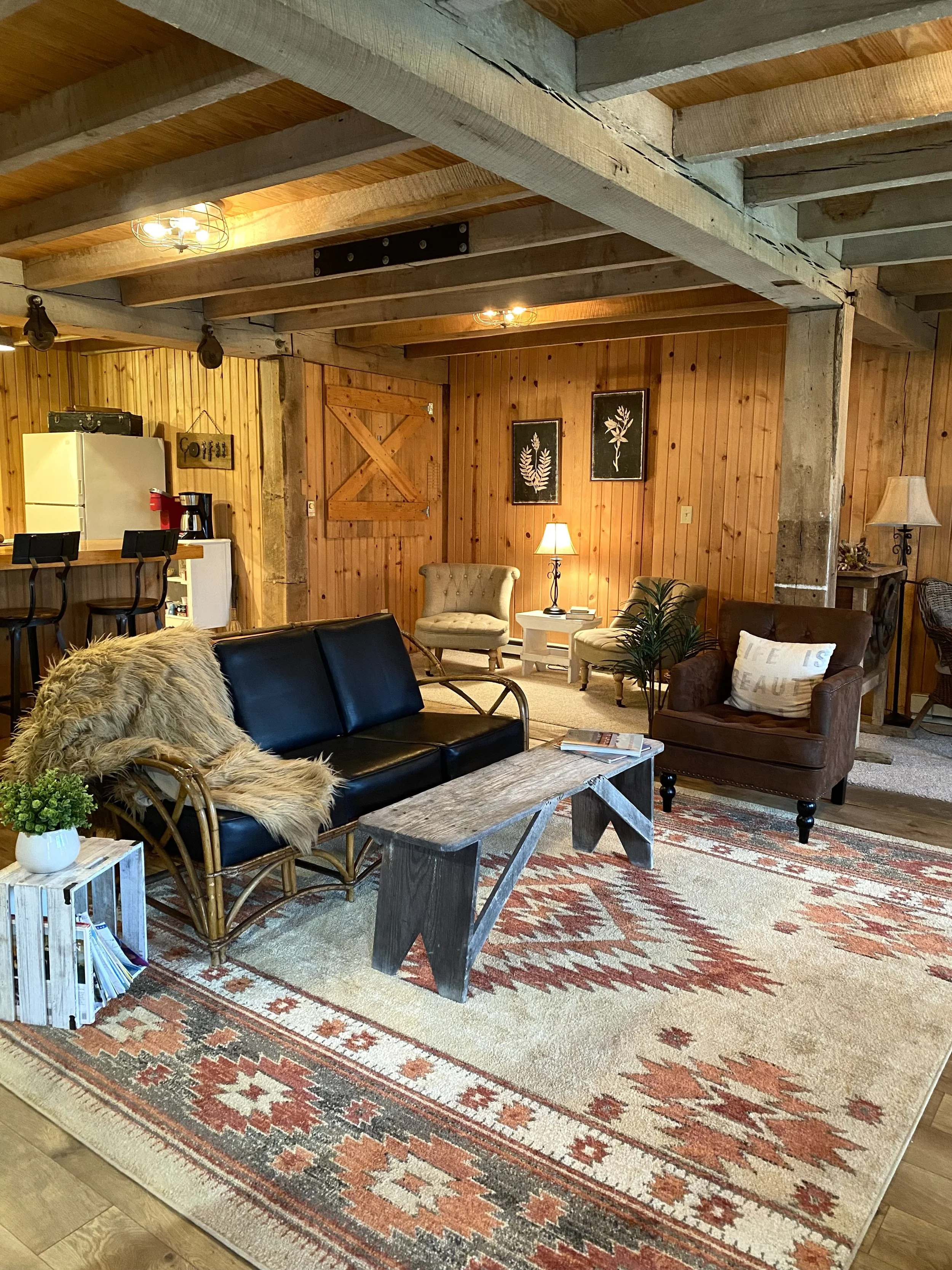 Cozy rustic living room with wood beams and paneling, featuring a black leather couch with a fur throw, a wooden coffee table, and an area rug with a geometric pattern. The room has a small white end table with books and a plant, a white mini fridge, and a coffee maker. Decorative elements include framed botanical art and a barn-style wooden door.