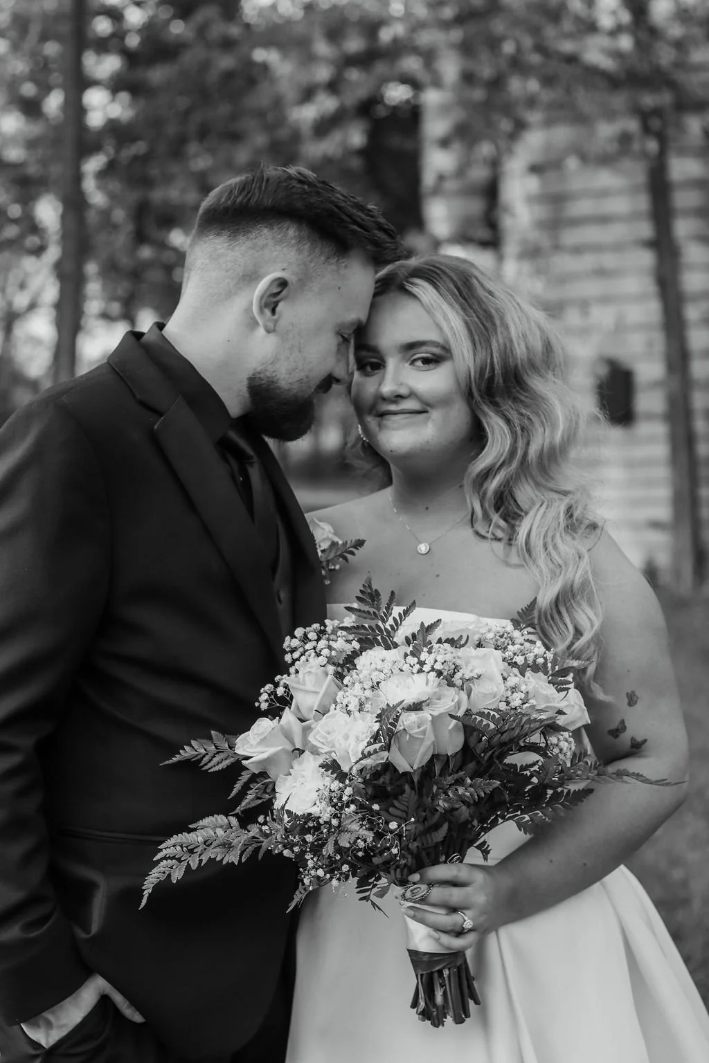 Black and white photo of a couple embracing; woman holds a bouquet of roses and foliage while man wears a suit.