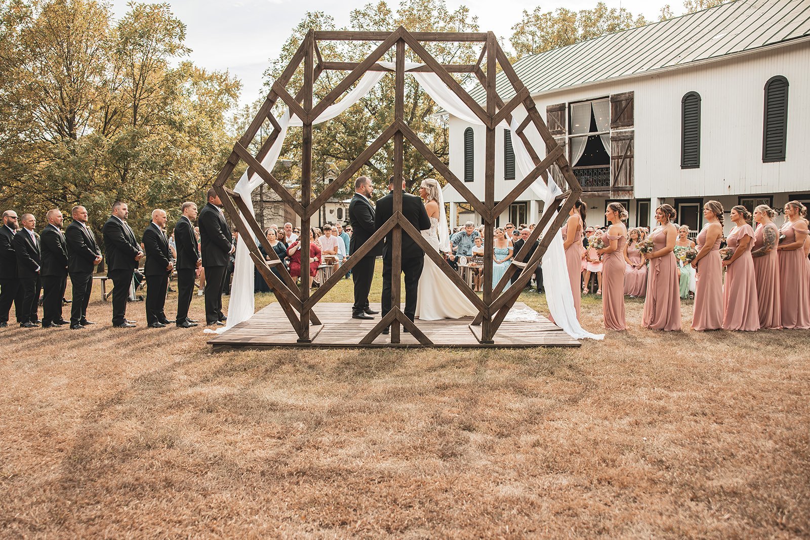 Outdoor wedding ceremony with couple exchanging vows under a wooden geometric arch adorned with white flowers and drapery. Bridesmaid and groomsman stand nearby, all dressed in formal attire. The Ohio Barn. Rustic Wedding Venue. Barn Venue. Ohio 