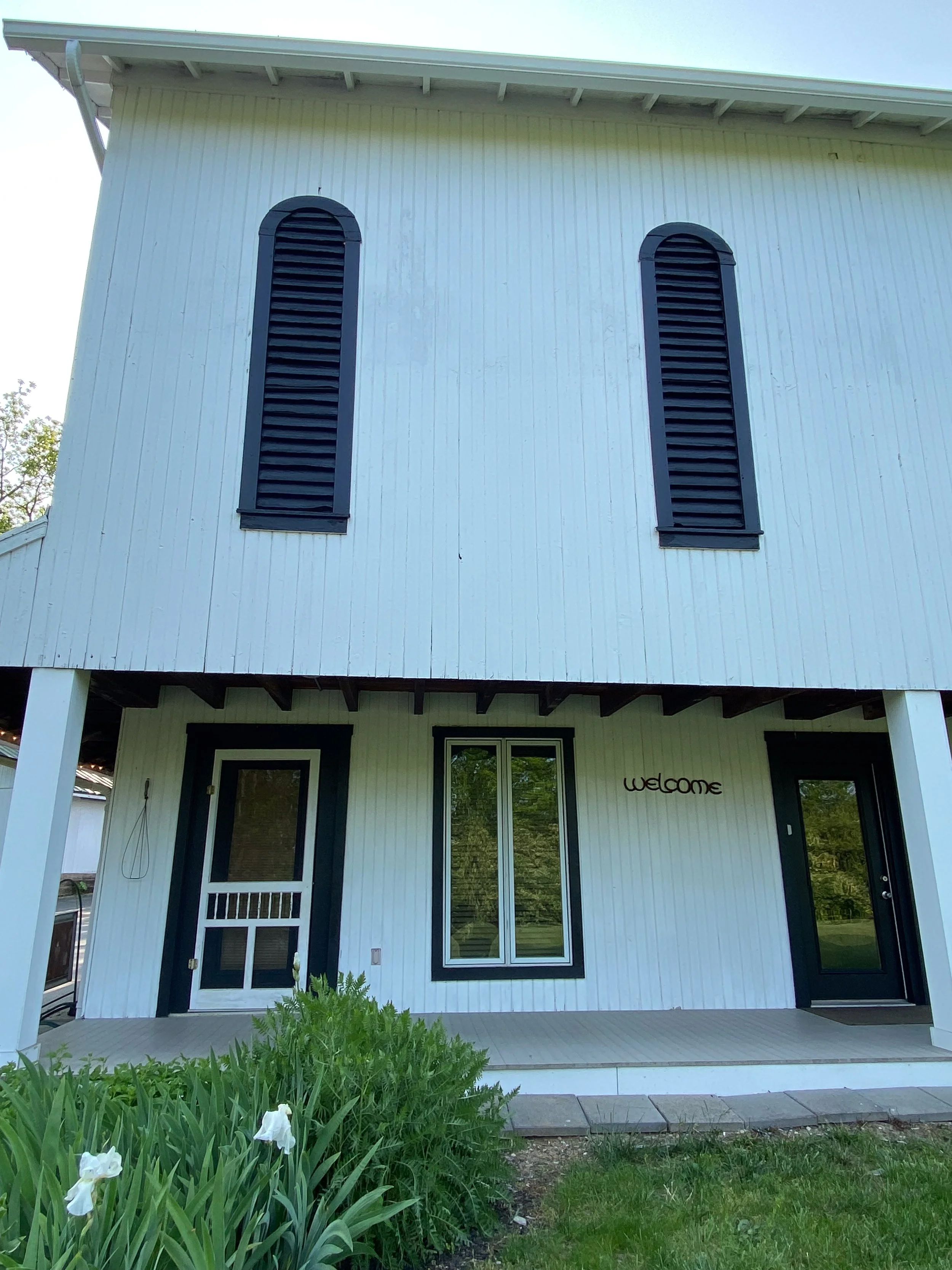 Two-story building exterior with white siding, two vertical louvered shutters, dark window frames, and a porch with a "Welcome" sign. The Ohio Barn. Yellow Springs, Ohio. Wedding Venue
