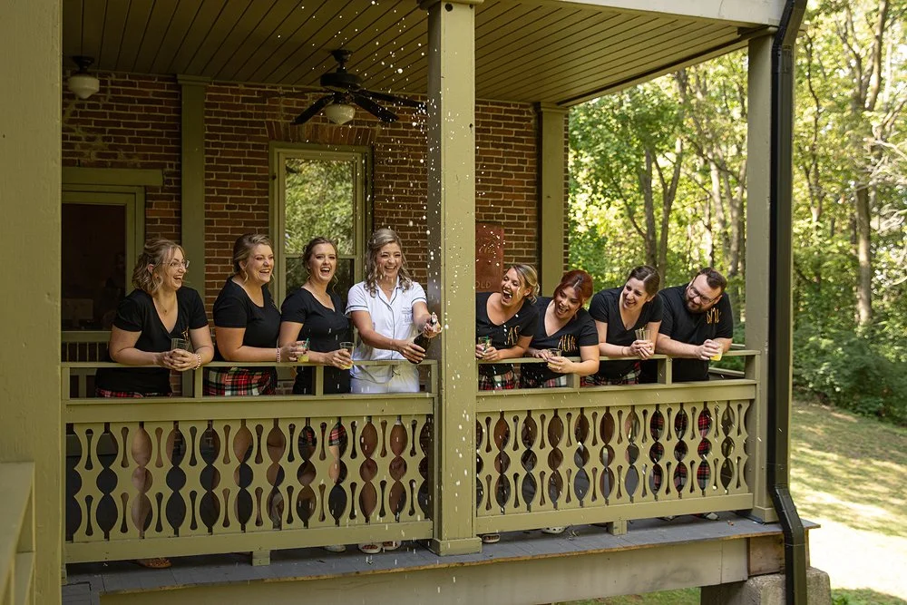 Group of people smiling and laughing on a porch, one person pouring a beverage, possibly champagne.