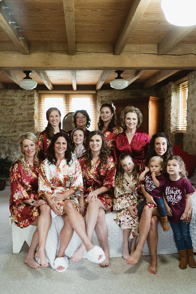 A group of women and young girls wearing floral and matching robes pose together indoors in a rustic room with wooden beams and stone walls.