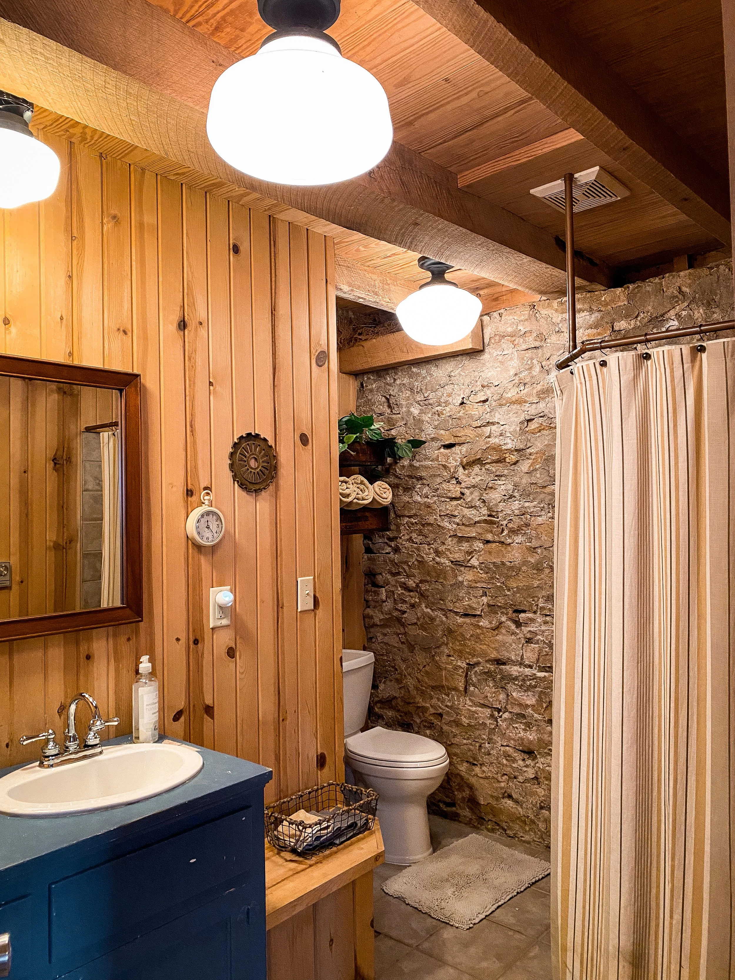 Rustic bathroom with wooden paneling, stone wall, and ceiling lamps. Features include a sink with a blue cabinet, a toilet, a wall clock, decorative shelf, and a shower with a curtain. Wedding Venue. Ohio. Southern Ohio