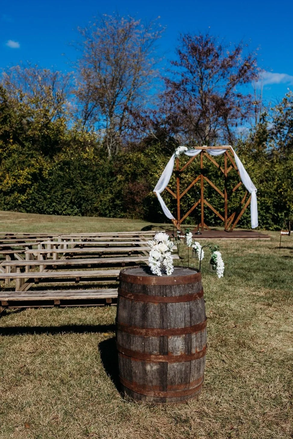 Outdoor wedding venue with wooden benches, a barrel with white flowers, and a geometric arch draped with fabric. Wedding Venue. Ohio Barn Venue. Fairborn, Ohio
