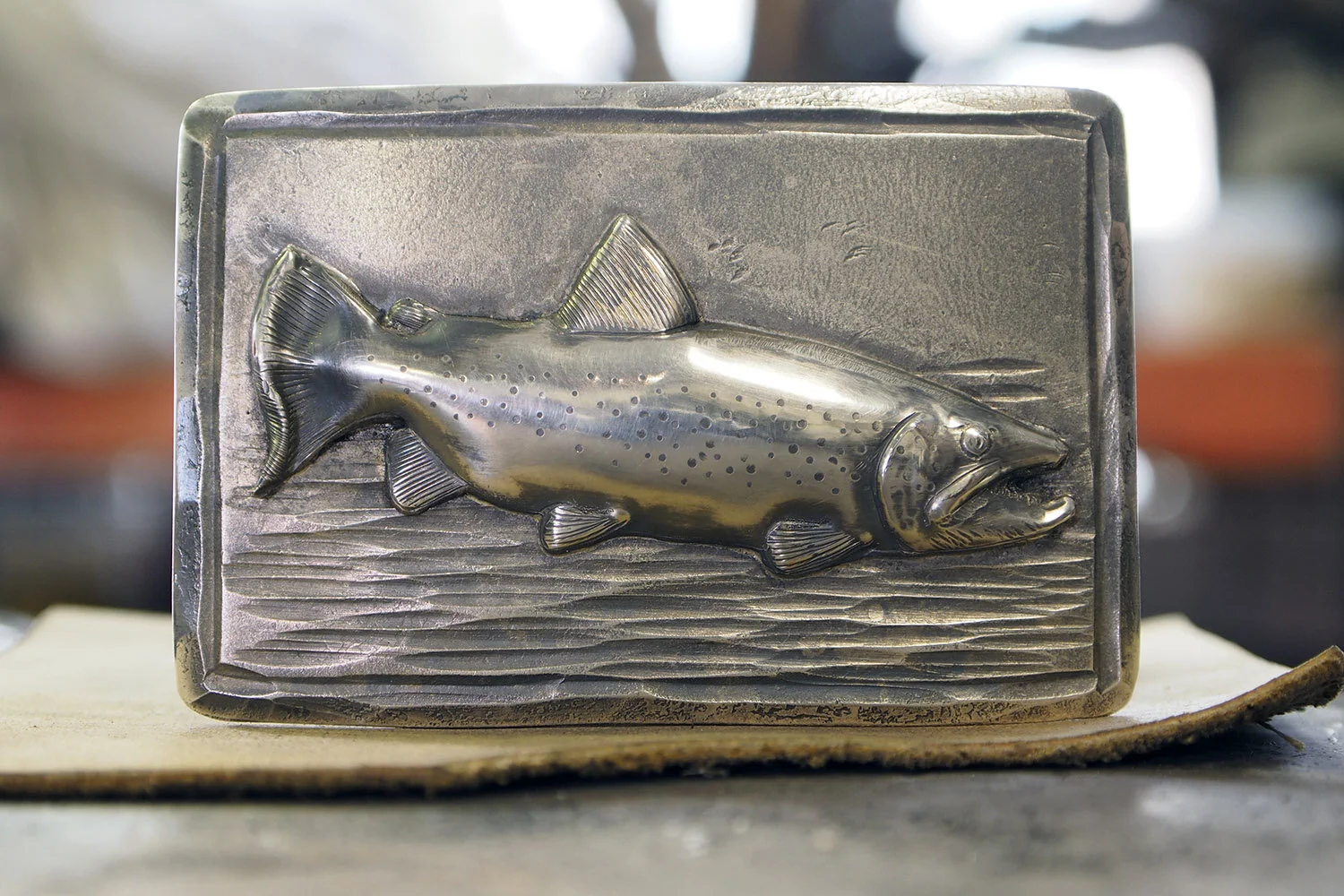 Brown trout in sterling silver on a 3" wide steel belt buckle. Forged water texture and chiseled border.