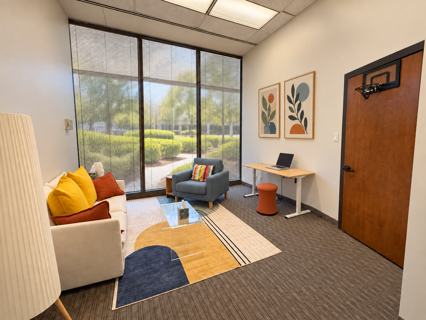 An office waiting area with a white sofa, yellow and rust-colored pillows, a blue armchair with a colorful pillow, a clear coffee table, a small wooden desk with a laptop, an orange stool, framed abstract art on the wall, large window with blinds, and a basketball hoop on the door.