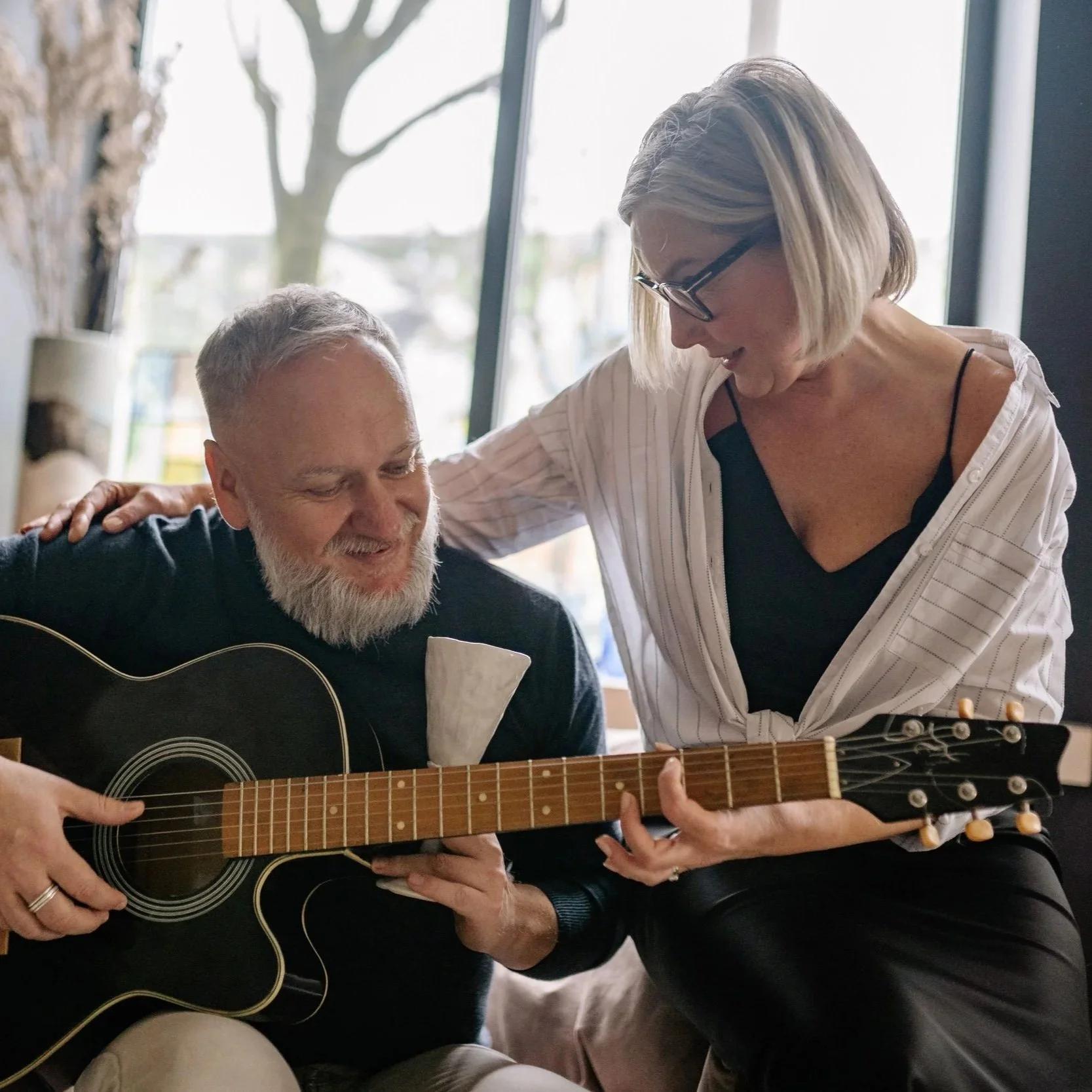 An older woman with blond hair and glasses helping an older man with a gray beard play a black acoustic guitar in a room with large windows and natural light.