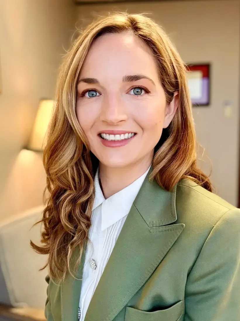 Photo of Dr. Jessica Klement, smiling, wearing a green blazer and white blouse, in an indoor setting with a lamp and framed artwork in the background.