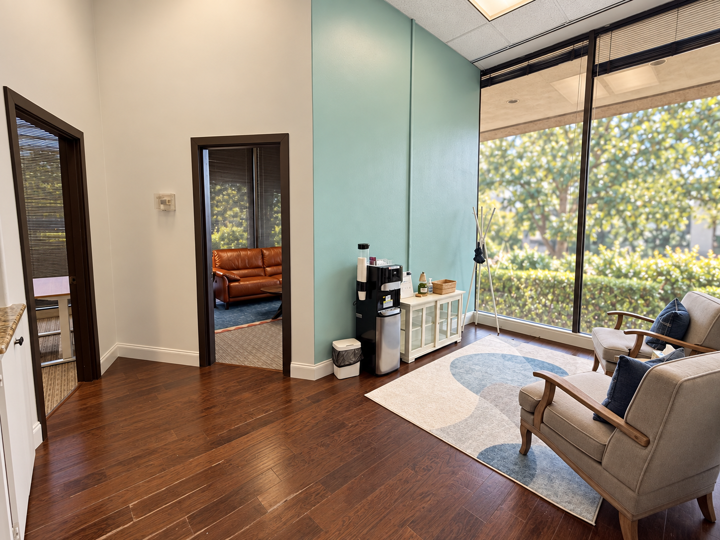 A waiting area with beige chairs, wooden armrests, and dark blue pillows. Large window with outside greenery, a small white table with a basket and bottles, and a coffee station with cups and supplies. An office doorway with viewing into another room with a brown leather sofa.