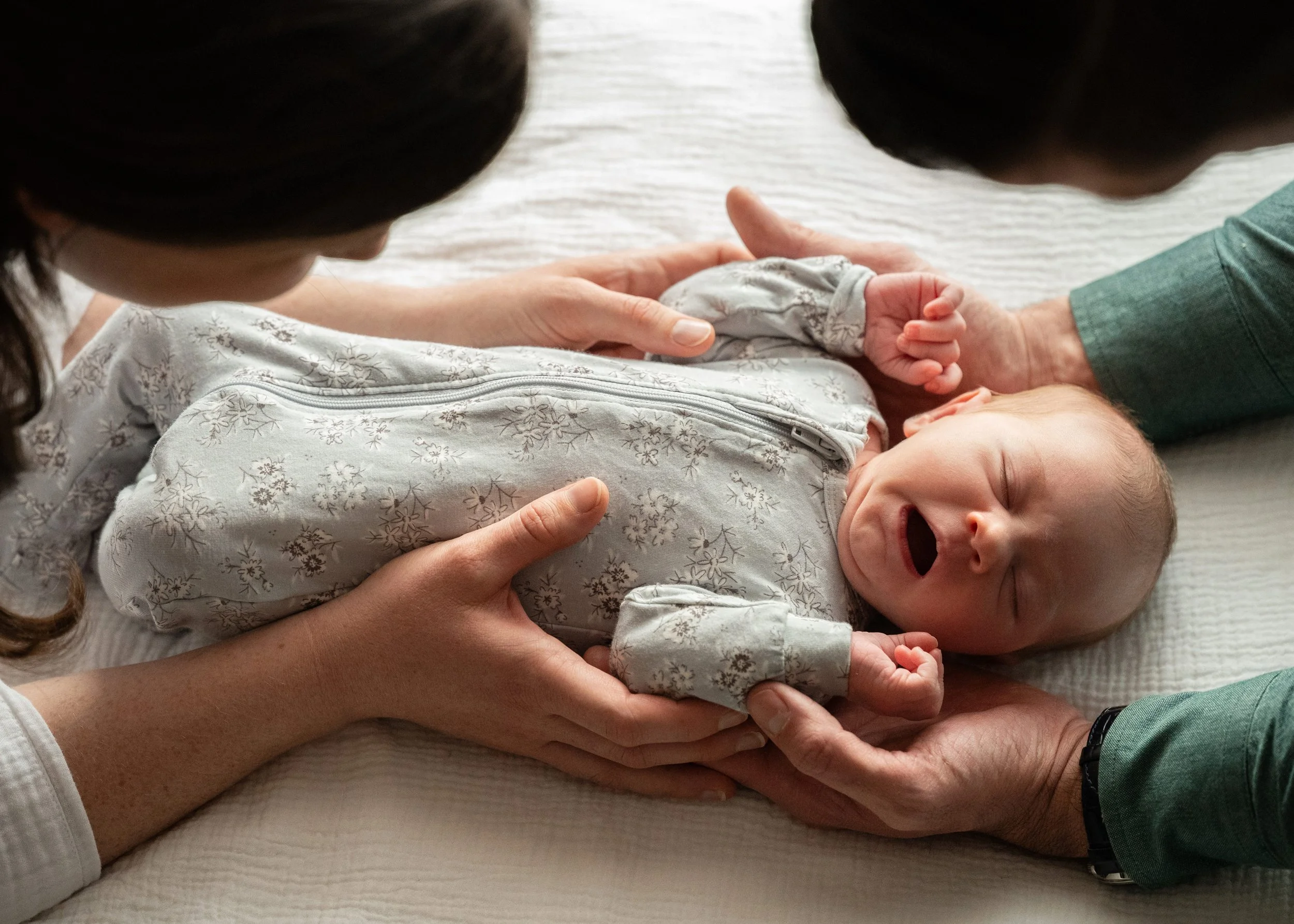 Newborn in grey sleeper laying on bed between parents looking from above with natural light