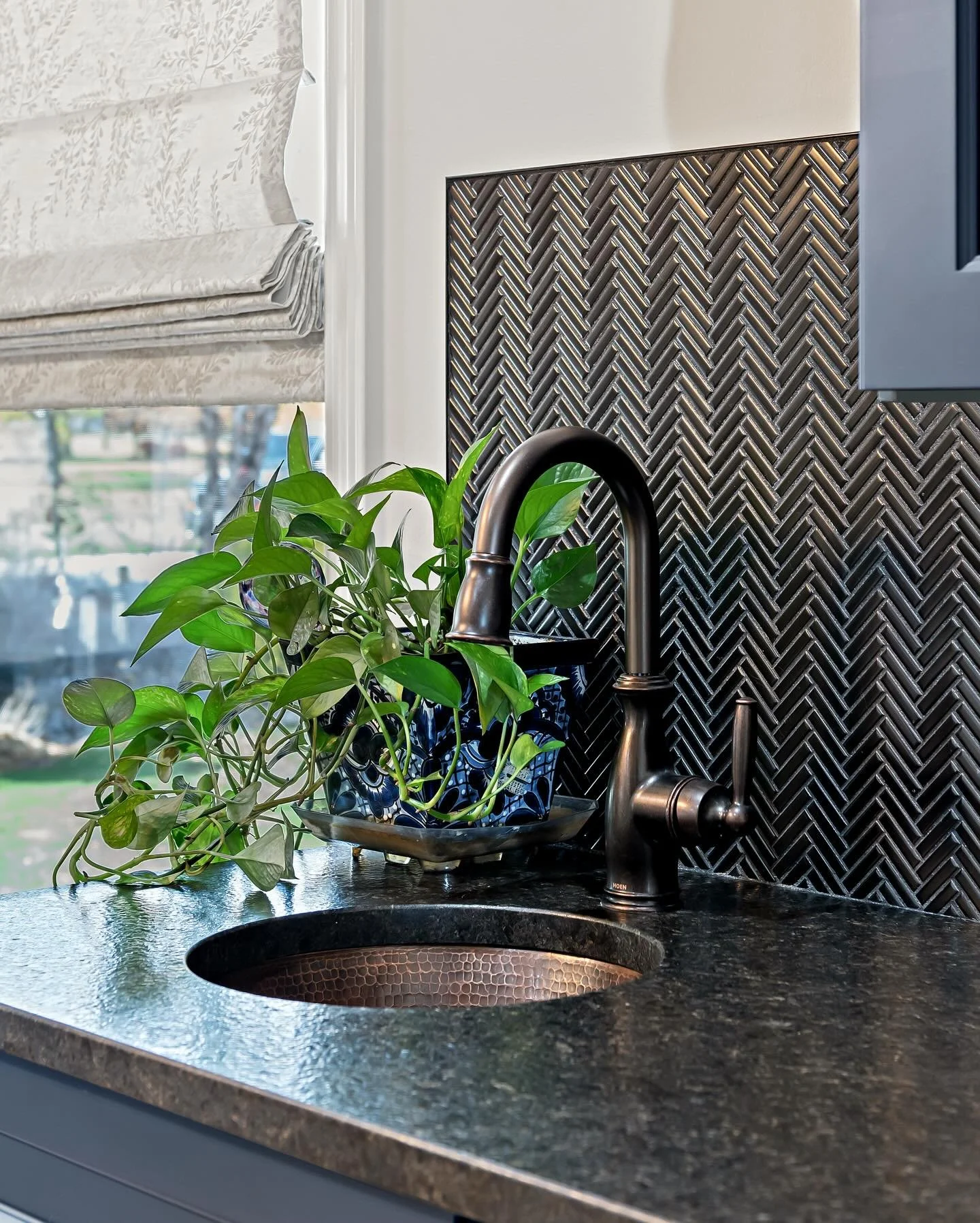 Love the details in this shot.
The black stone herringbone backsplash sets the mood, the bronze faucet and copper sink add warmth, and the pop of green from the plant brings it all to life. Finished with a soft Roman shade and just a peek of blue cab
