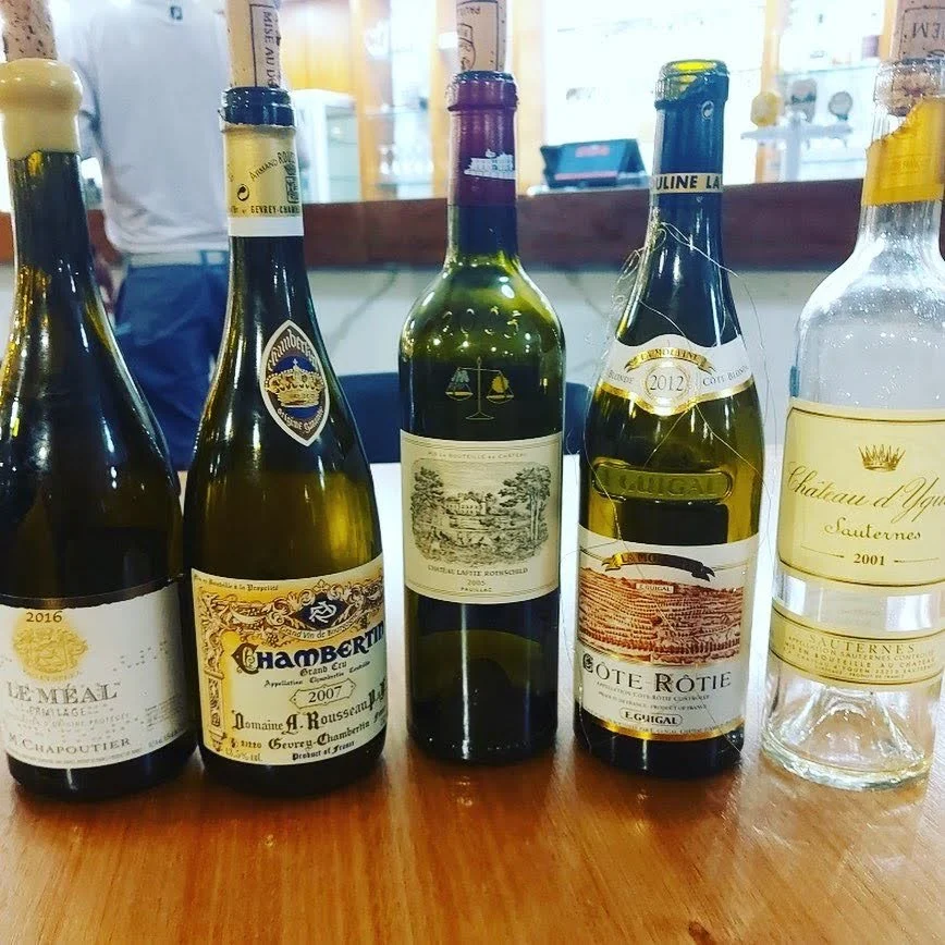 Five bottles of white wine lined up on a wooden table in a restaurant or café setting.