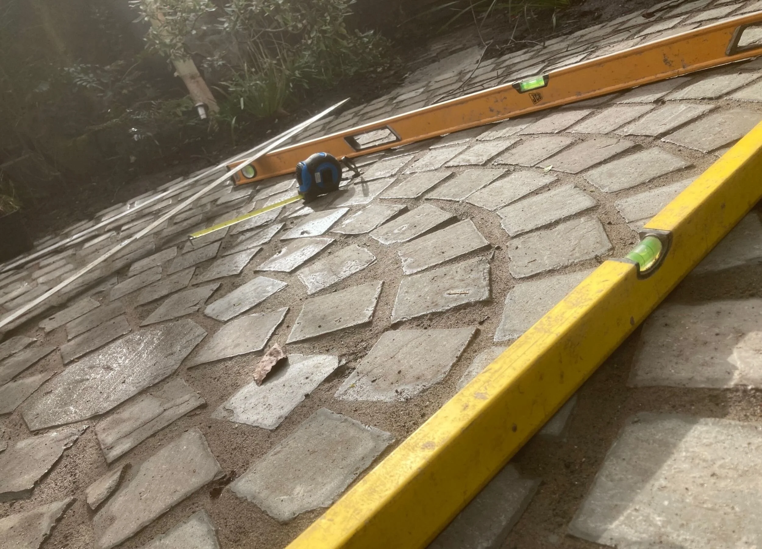 Construction tools including a yellow level, a tape measure, and a orange level are placed on a brick-tiled surface, with some greenery in the background.