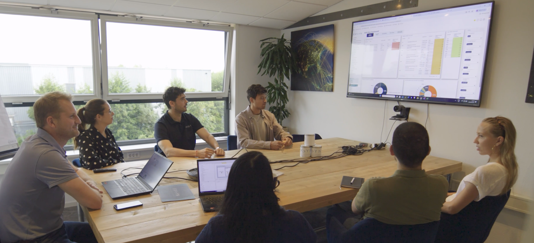 People sitting around a conference table in a meeting room, watching a presentation on a large screen displaying data charts and spreadsheets.