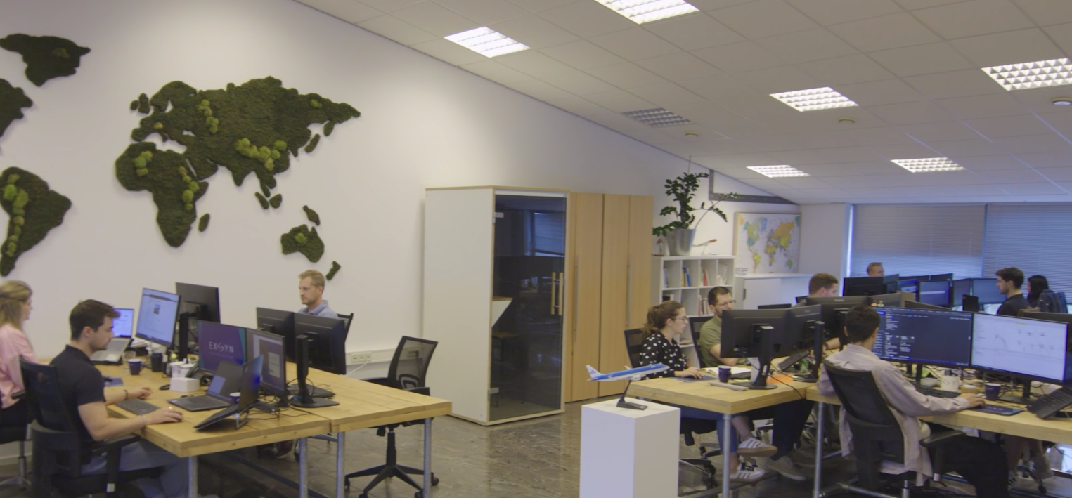 An office space with multiple people working at their desks with computer monitors. The room has a world map on the wall, a globe, and plants, with bright overhead lighting and window blinds.