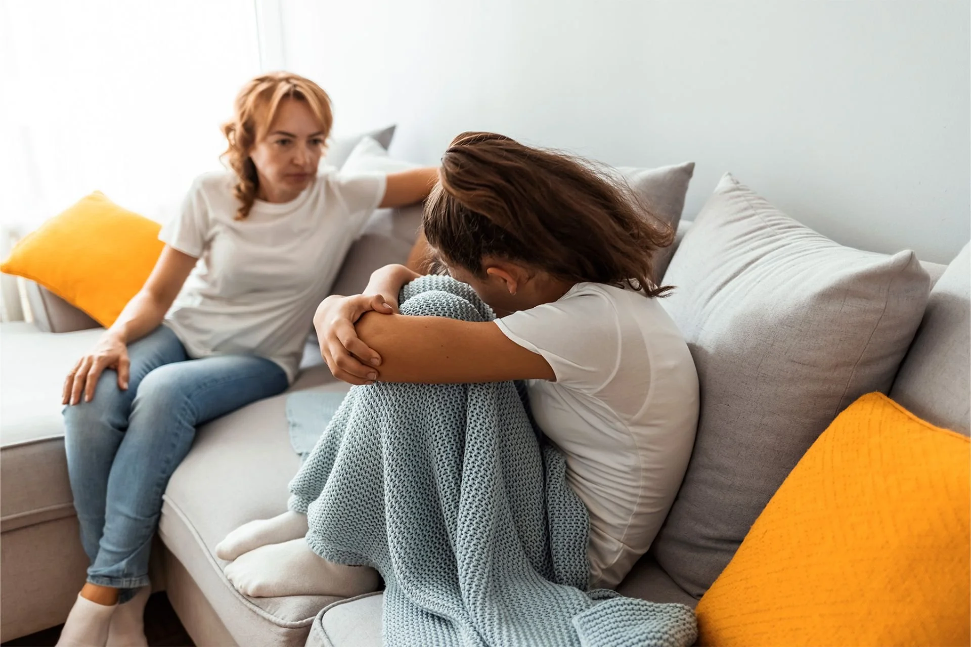A girl appearing distressed sits on a couch during a therapy session, talking with a therapist who is holding a notebook and gesturing with her hand.