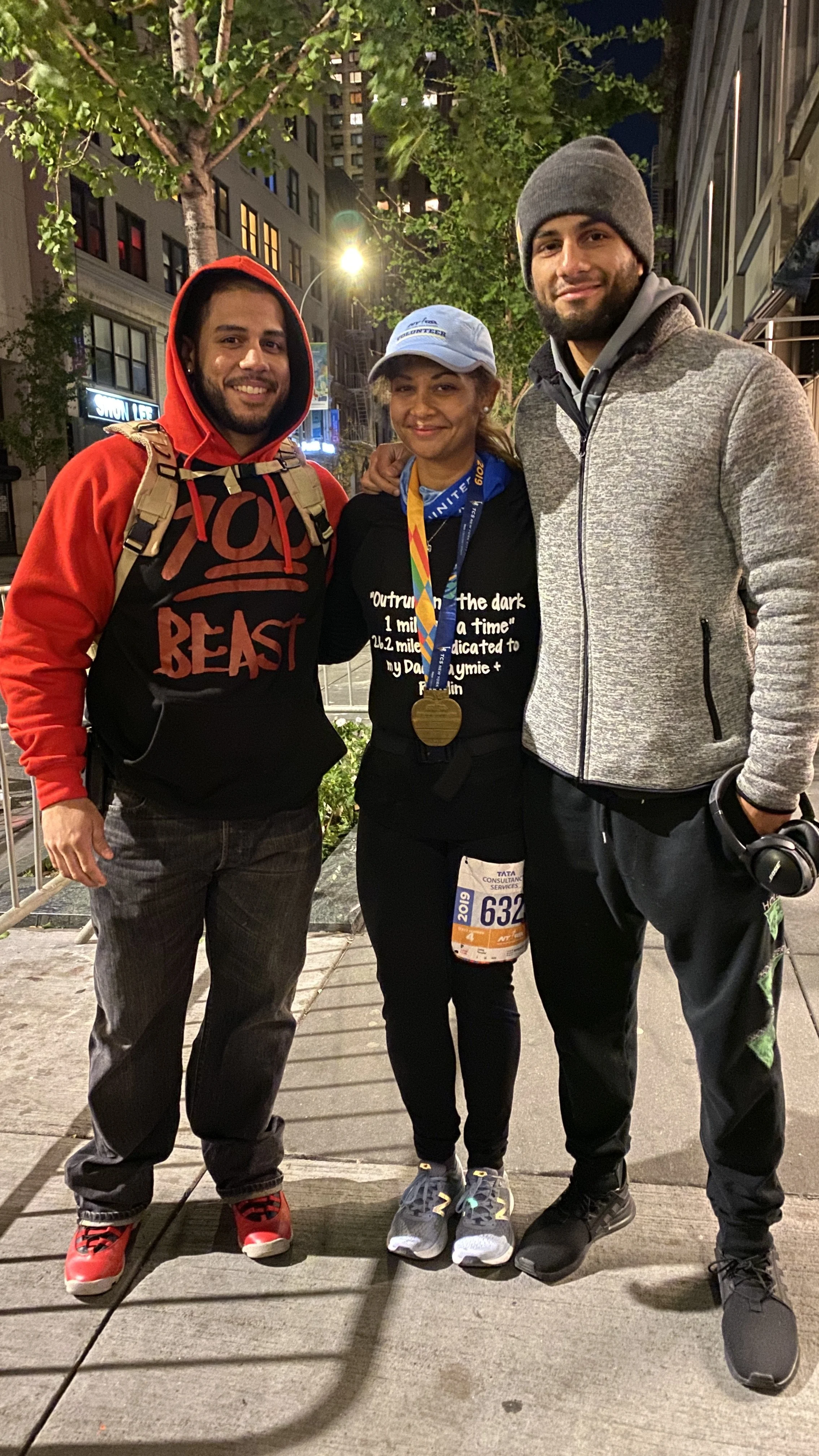 Three people standing together outdoors at night, including a woman in race gear with a medal and two men in casual clothing, smiling for the camera.