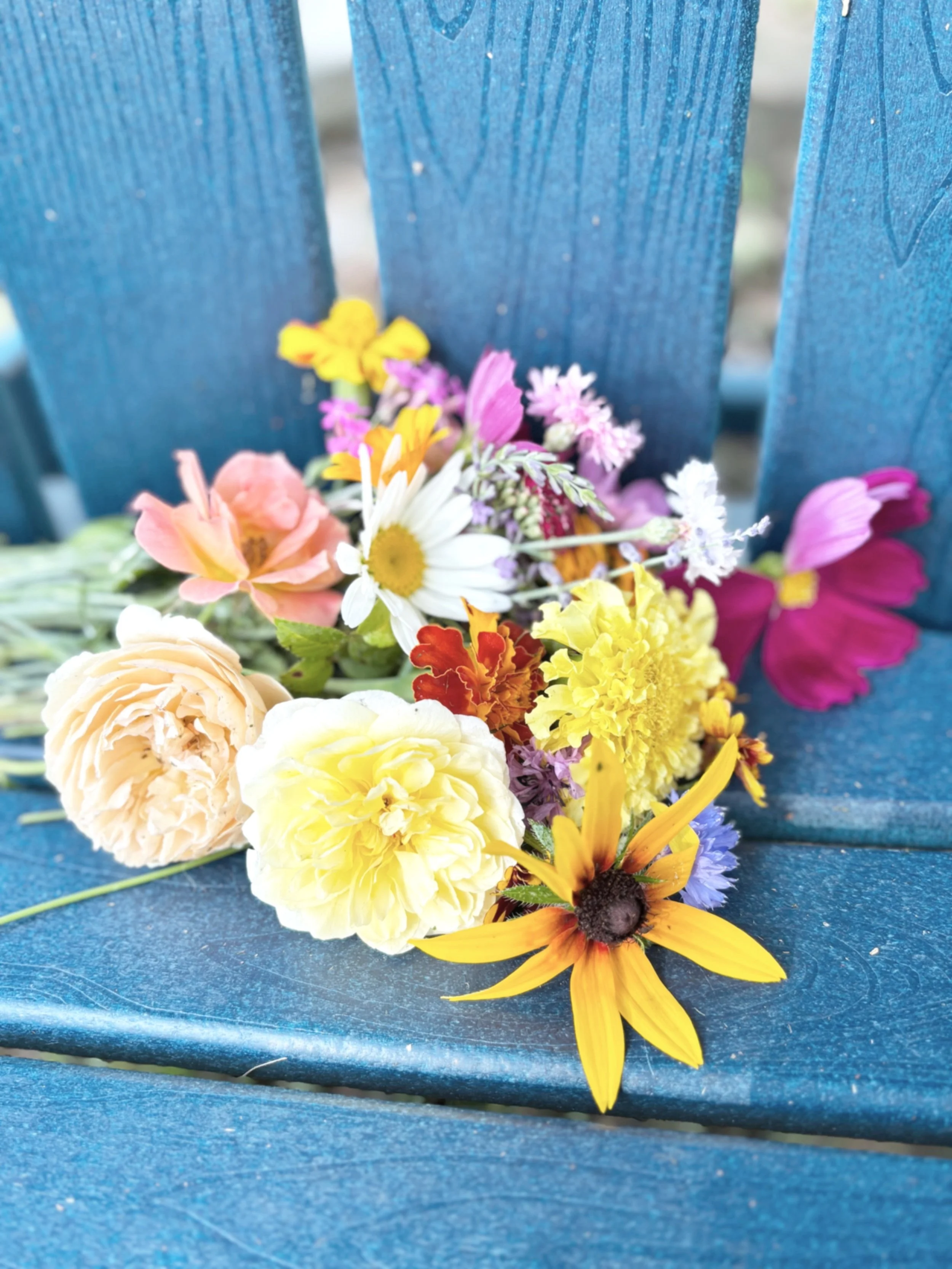 A colorful bouquet of various flowers including daisies, sunflowers, and roses, placed on a blue wooden outdoor chair.