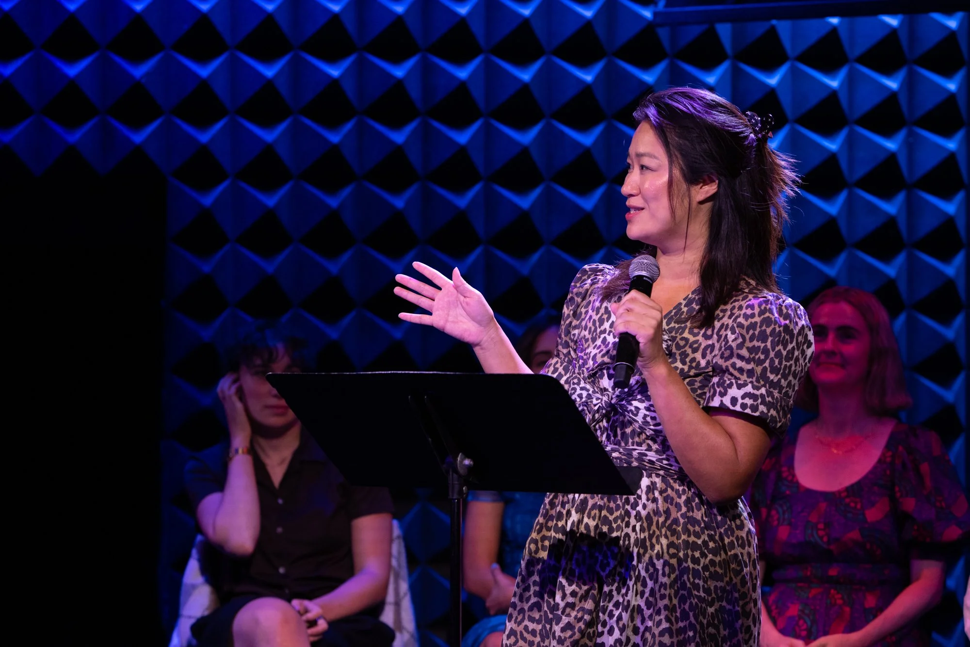 A woman with long dark hair, wearing a leopard print dress, speaking into a microphone on stage with a blue geometric background, as an audience watches.