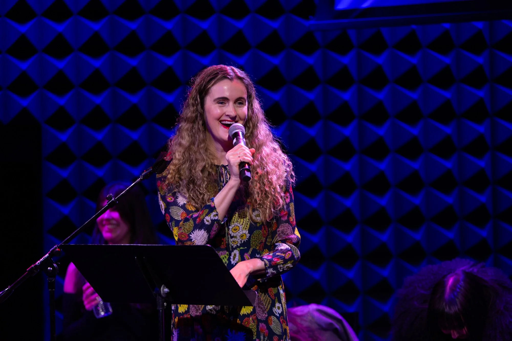 A woman with long curly hair wearing a colorful floral dress is smiling and holding a microphone on stage with a blue geometric background.