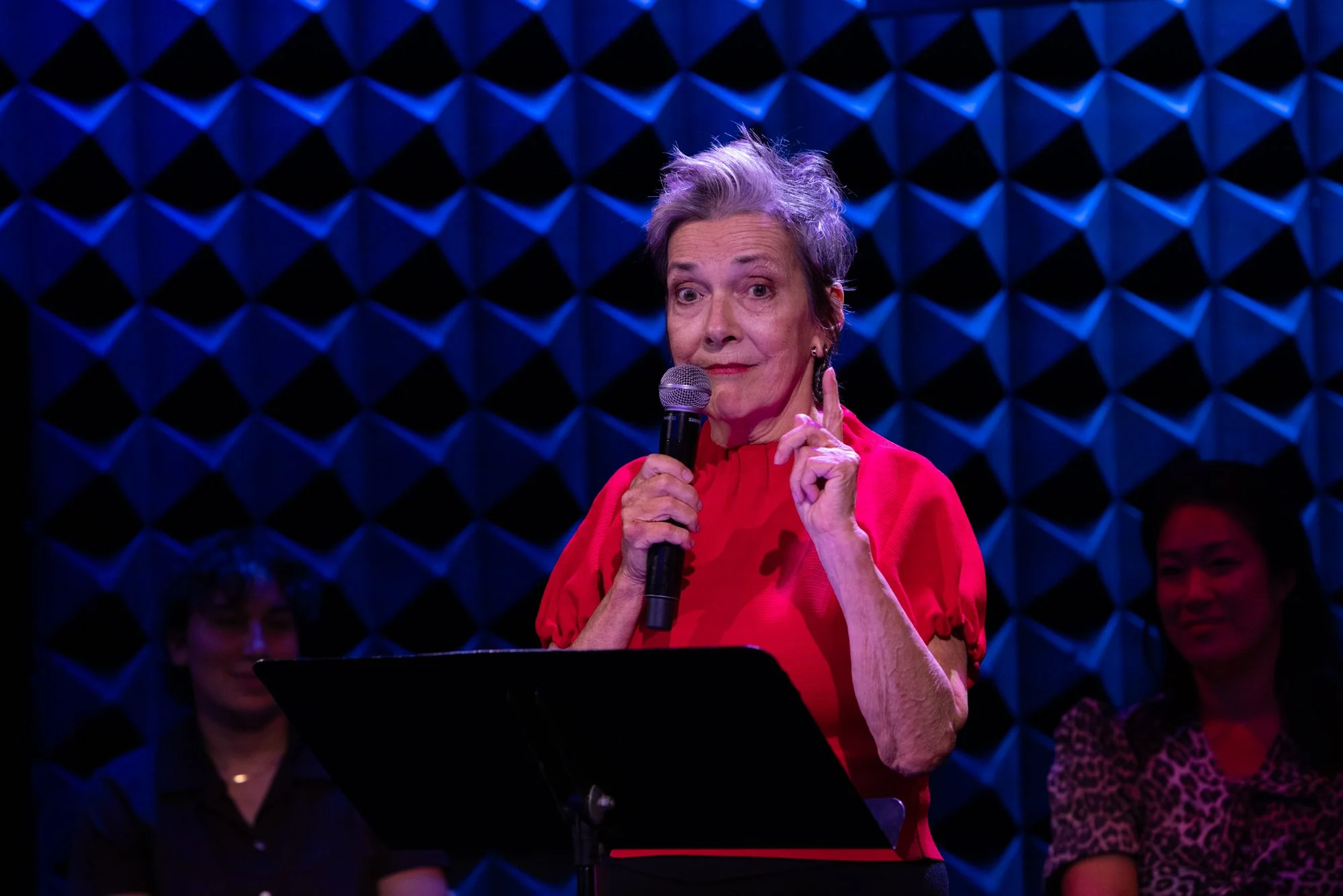 An elderly woman with short, gray hair speaking into a microphone while wearing a red blouse, standing in front of a dark blue, diamond-patterned acoustic wall, with several women seated behind her.