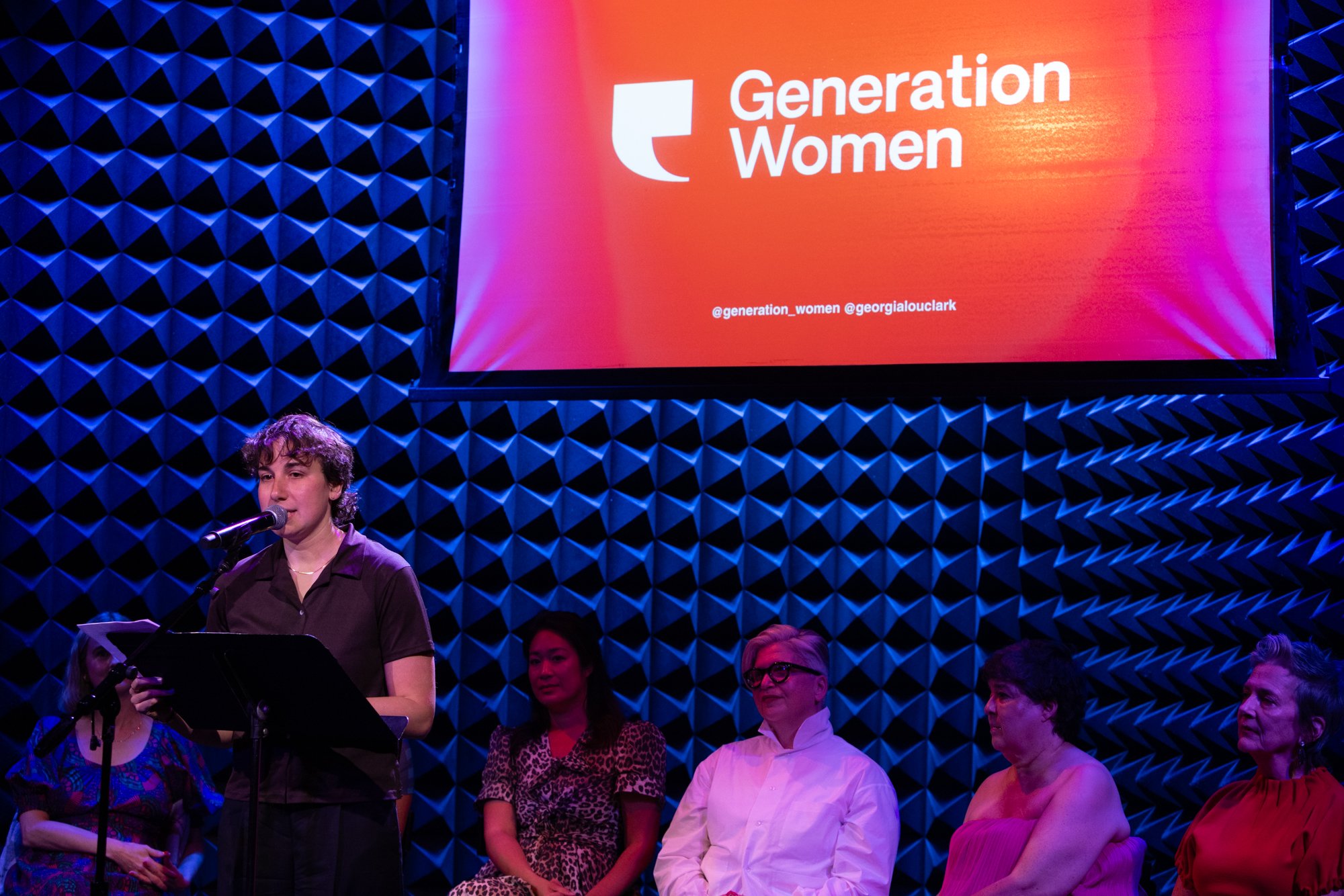 A woman speaking into a microphone while holding a paper at a Generation Women event, with a large red and orange screen displaying the organization's name and social media handles behind her, and several women seated and listening in a room with blu
